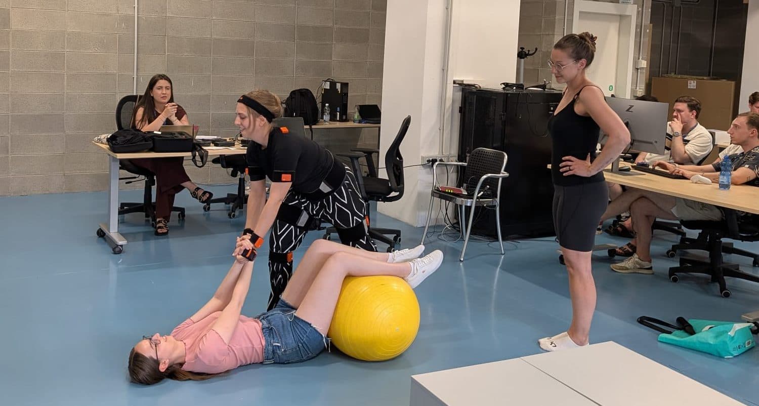 Eine Frau hilft einer anderen Frau beim Dehnen, während diese in einem modernen Büro auf einem gelben Gymnastikball liegt. Im Hintergrund sitzen mehrere Personen an Schreibtischen und beobachten das Geschehen.