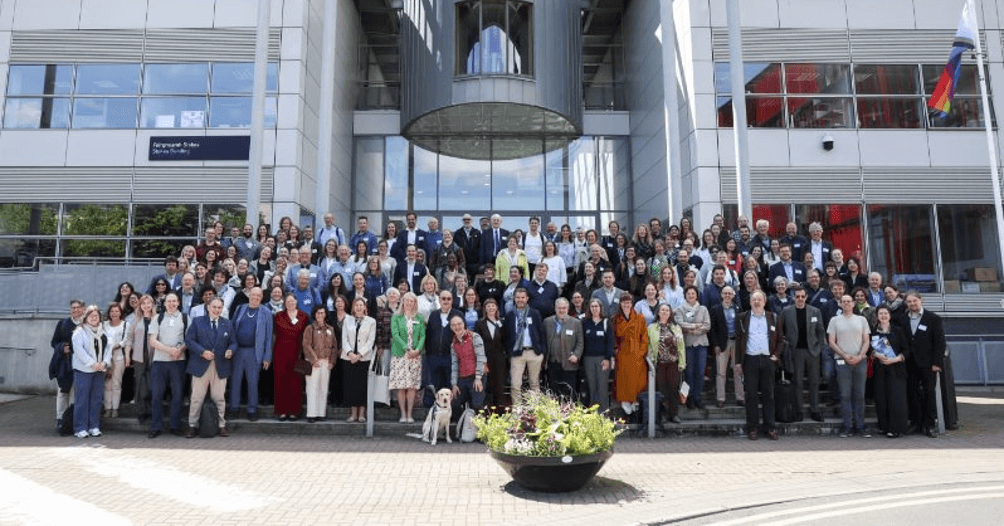 Eine große Gruppe von Menschen steht auf den Stufen vor einem modernen Gebäude und posiert für ein Gruppenfoto. In der ersten Reihe steht ein Blindenhund, und im Vordergrund ist ein Blumenkübel zu sehen.