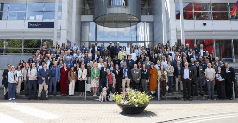 Eine große Gruppe von Menschen posiert für ein Gruppenfoto auf den Stufen vor einem modernen Bürogebäude; im Vordergrund sind ein Blumenkübel und ein Blindenhund zu sehen.