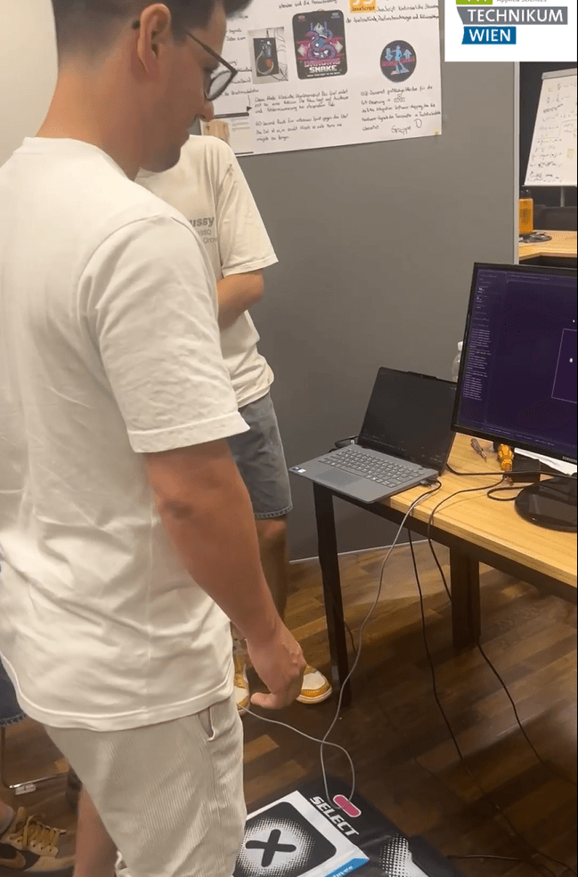Eine Person steht auf einem Tanzpad vor einem Schreibtisch, auf dem ein Laptop und ein Monitor stehen, auf dem Code angezeigt wird, während eine andere Person zusieht. Beide befinden sich in einem Klassenzimmer oder Labor, an dessen Wänden Plakate hängen.