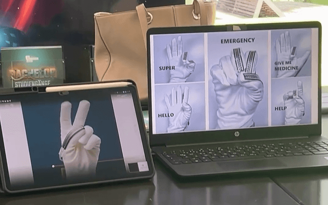 Auf einem Tablet und einem Laptop sind Bilder einer Hand in einem weißen Handschuh zu sehen, die verschiedene Gesten macht. Auf dem Laptop-Bildschirm sind die Gesten mit den Begriffen „NOTFALL“, „GIB MIR MEDIKAMENTE“, „HILFE“, „HALLO“ und „SUPER“ beschriftet. Im Hintergrund ist eine beige Handtasche zu sehen.