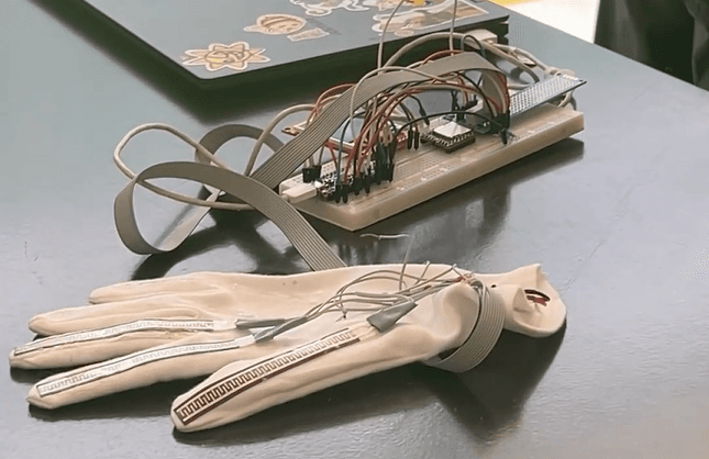 Ein weißer Handschuh, an dem Drähte und Sensoren befestigt sind, ist mit einem elektronischen Steckbrett verbunden, auf dem verschiedene Bauteile und Kabel angebracht sind. Im Hintergrund ist teilweise ein Laptop mit Aufklebern zu sehen.