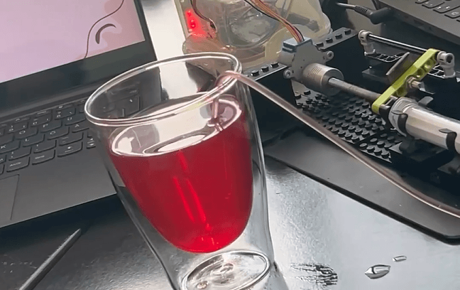 Ein mit roter Flüssigkeit gefülltes Glas ist mit einem Schlauch verbunden, der zu einem mechanischen Gerät auf einem Schreibtisch in der Nähe eines Laptops führt. Auf der Oberfläche des Schreibtisches sind einige Wassertropfen zu sehen.