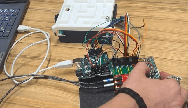 Eine Hand justiert Kabel, die mit einem Arduino-Board und anderen elektronischen Bauteilen auf einem Tisch verbunden sind; daneben stehen ein Laptop und ein weißes Labyrinthgerät aus Kunststoff.