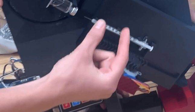Eine Hand zeigt auf eine Spritze, die an einem schwarzen mechanischen Gerät befestigt ist, wobei im Hintergrund Kabel und Bauteile zu sehen sind.