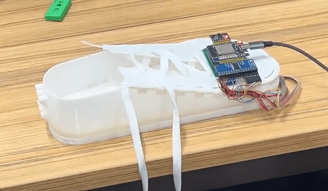 Auf einem Holztisch steht ein weißes Kunststoffgerät mit Riemen und einer daran befestigten elektronischen Leiterplatte, einschließlich Drähten und einem Stromkabel.