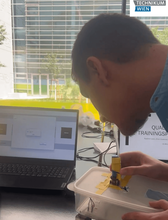 Eine Person betrachtet aufmerksam ein kleines elektronisches Gerät, das an einer durchsichtigen Plastikbox befestigt ist; im Hintergrund ist ein Laptop zu sehen, auf dem eine Software läuft und das Logo „Technikum Wien“ zu erkennen ist.