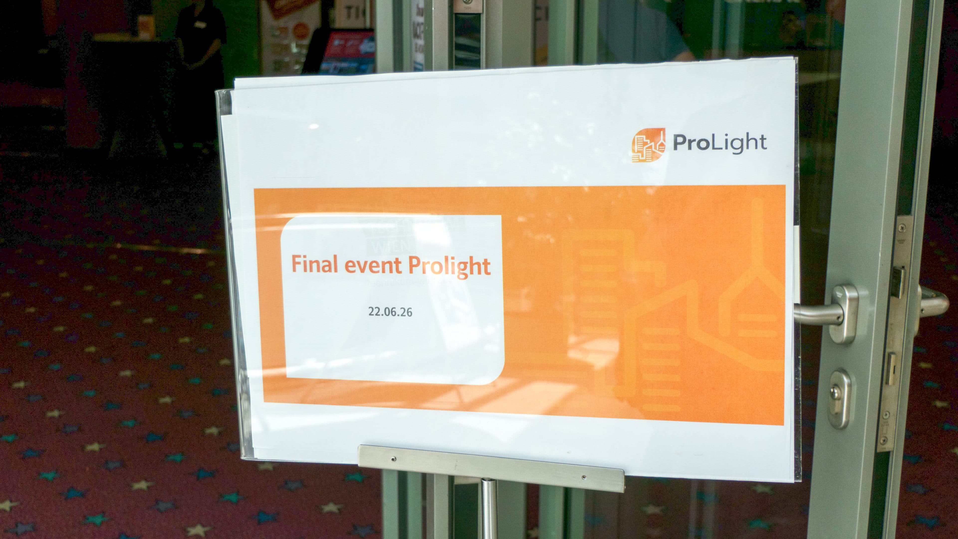 Auf einem Schild an einer Glastür ist das ProLight-Logo zu sehen, dazu die Aufschrift „Final event Prolight“ mit dem Datum „22.06.26“ auf orangefarbenem Hintergrund. Die Tür steht einen Spalt breit offen und gibt den Blick auf einen gemusterten Teppichboden im Inneren frei.