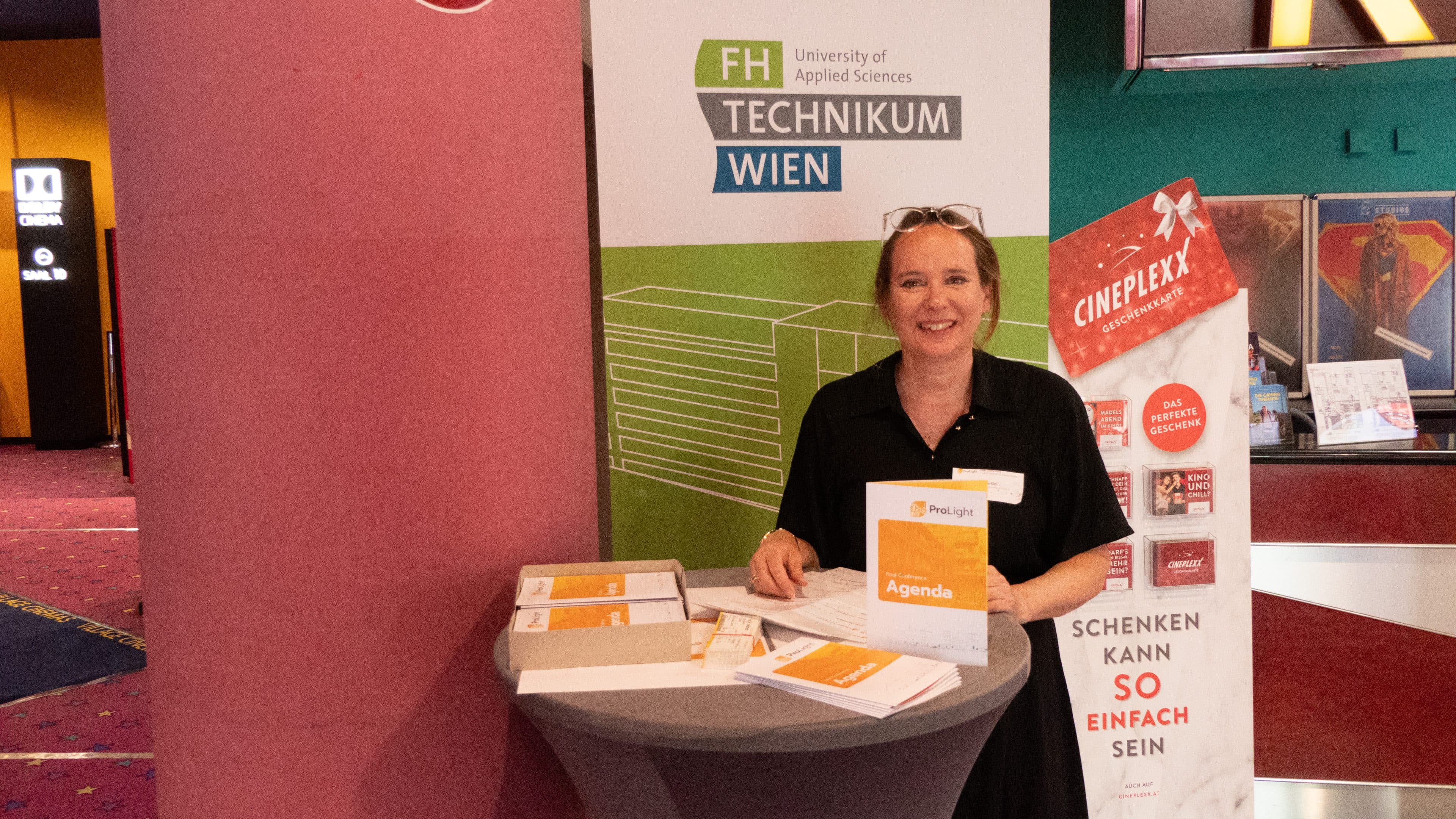 Bei einer Veranstaltung steht eine Frau hinter einem Tisch mit Broschüren und lächelt in die Kamera. Hinter ihr sind ein Banner der Fachhochschule Technikum Wien und ein Cineplexx-Plakat zu sehen.