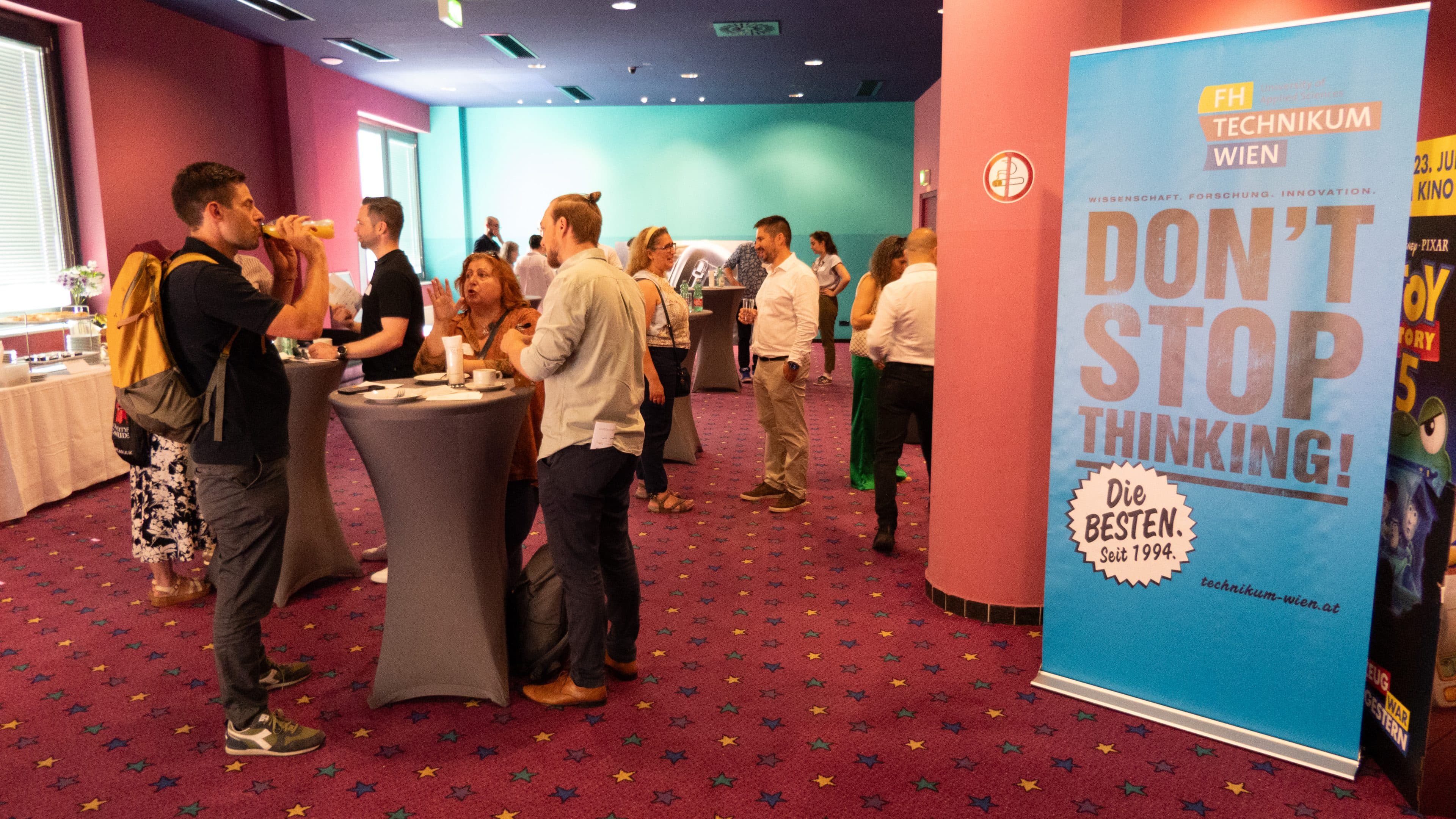 Menschen, die sich an hohen Tischen in einem hellen, mit Teppichboden ausgelegten Konferenzraum unterhalten und Kontakte knüpfen. Auf einem großen blauen Schild steht: „DONT STOP THINKING! Die BESTEN. Seit 1994.“ Das Logo der FH Technikum Wien ist zu sehen.