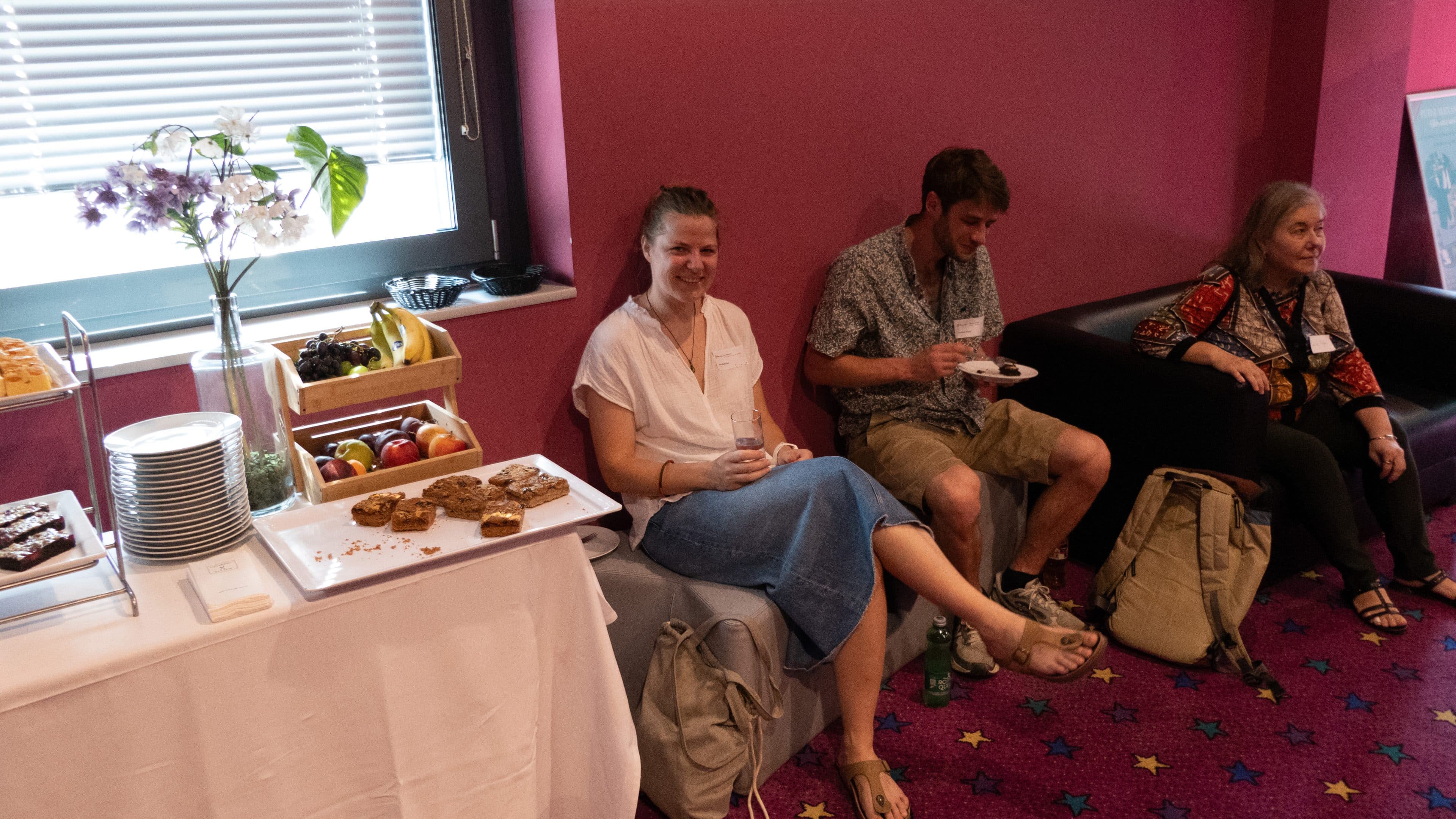Drei Personen sitzen in einem hell erleuchteten Raum mit rosa Wänden und einem lila Teppich mit Sternenmuster und essen Snacks; neben ihnen steht ein Tisch mit Tellern, Snacks und Obst, und auf der Fensterbank steht eine Blumenvase.
