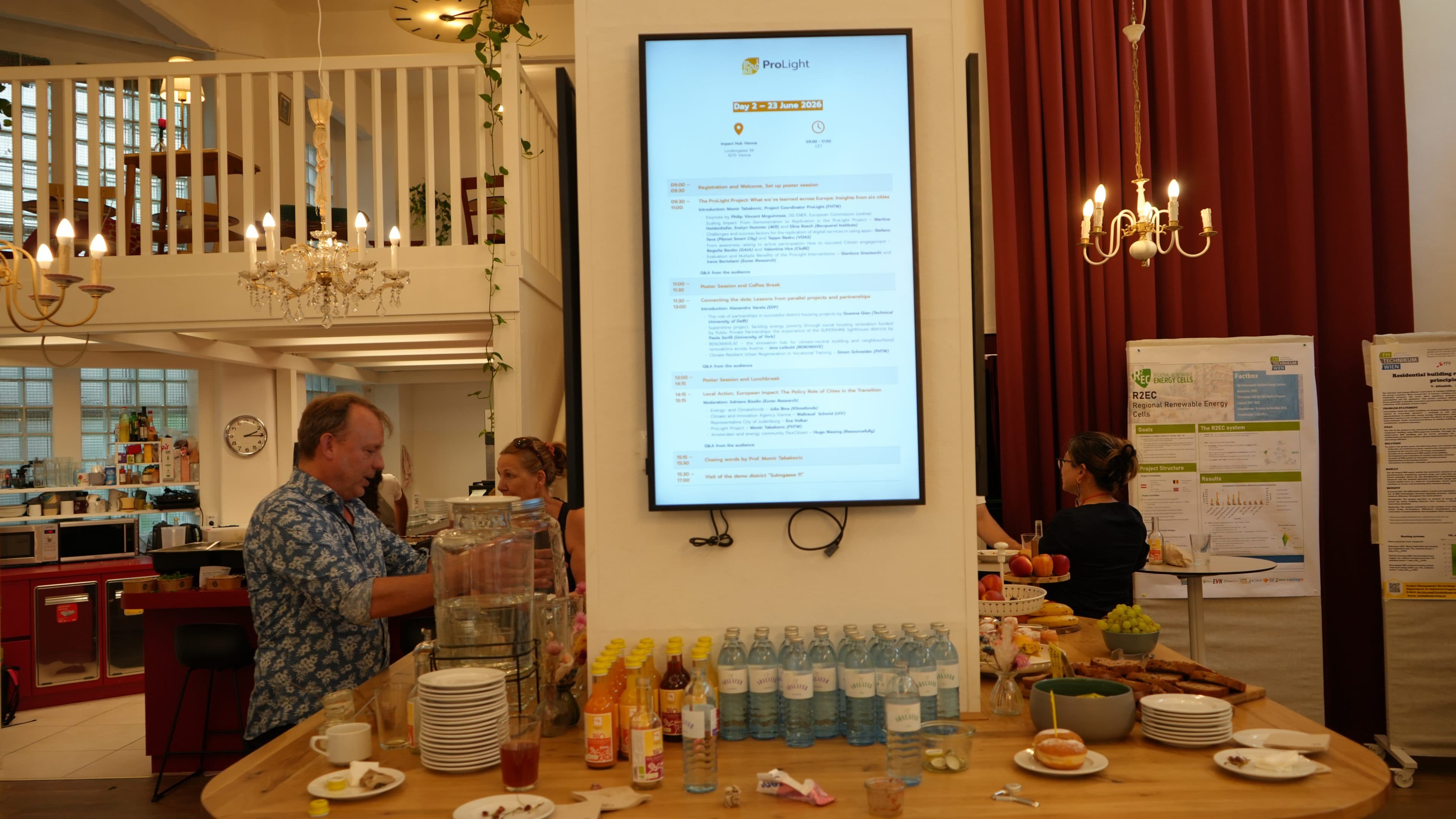 In einem hellen Raum steht ein Tisch mit Getränken, Obst und Snacks. Die Gäste bedienen sich selbst. An der Wand hängt ein großer Bildschirm, auf dem das Veranstaltungsprogramm angezeigt wird. Kronleuchter und rote Vorhänge runden die Einrichtung ab.