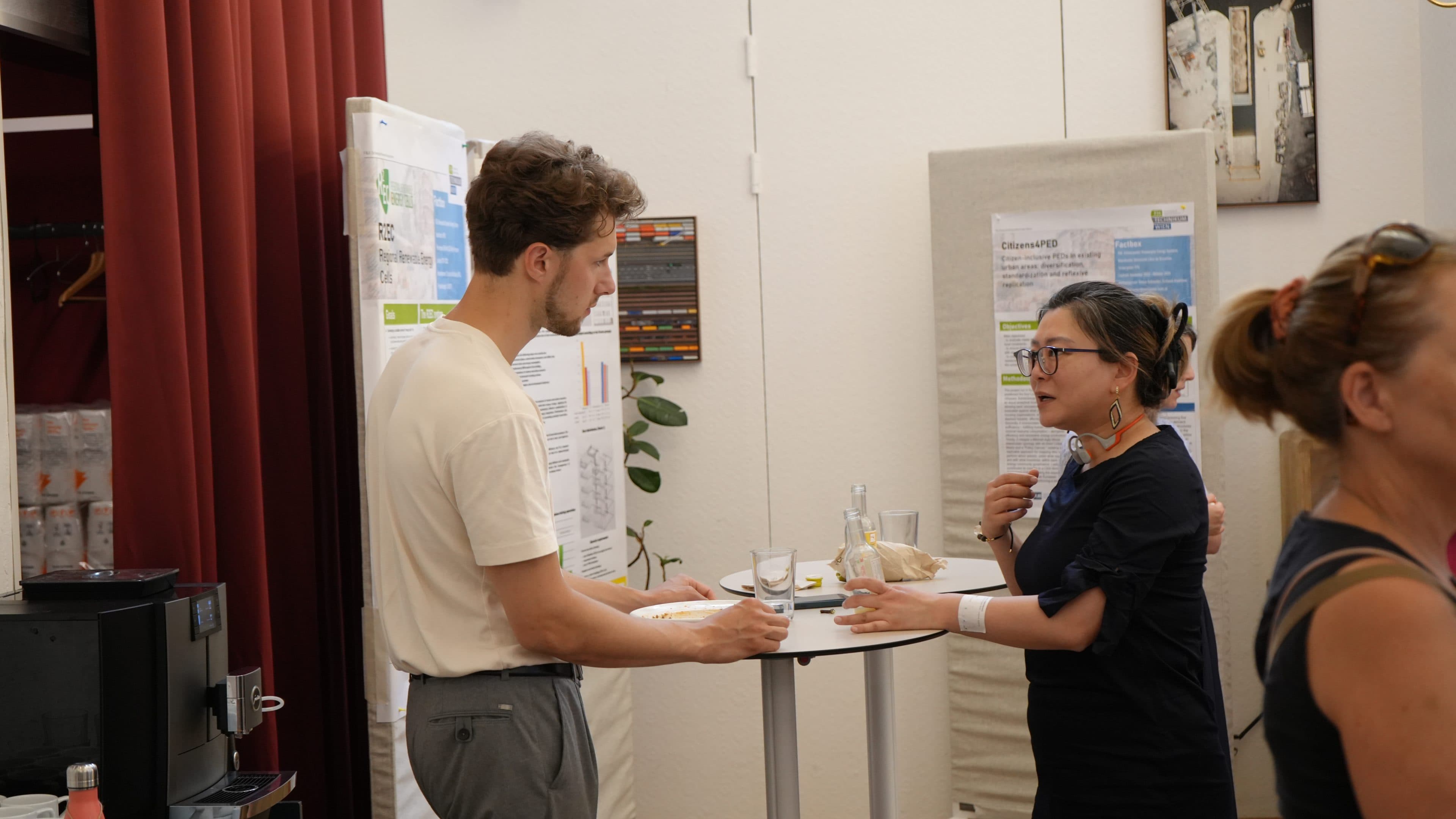 Zwei Personen stehen an einem kleinen runden Tisch in einem Raum, in dessen Nähe Plakate und Präsentationswände zu sehen sind, und unterhalten sich bei einem Getränk. Eine Person gestikuliert beim Sprechen, die andere hört zu. Rechts ist eine dritte Person teilweise zu sehen.