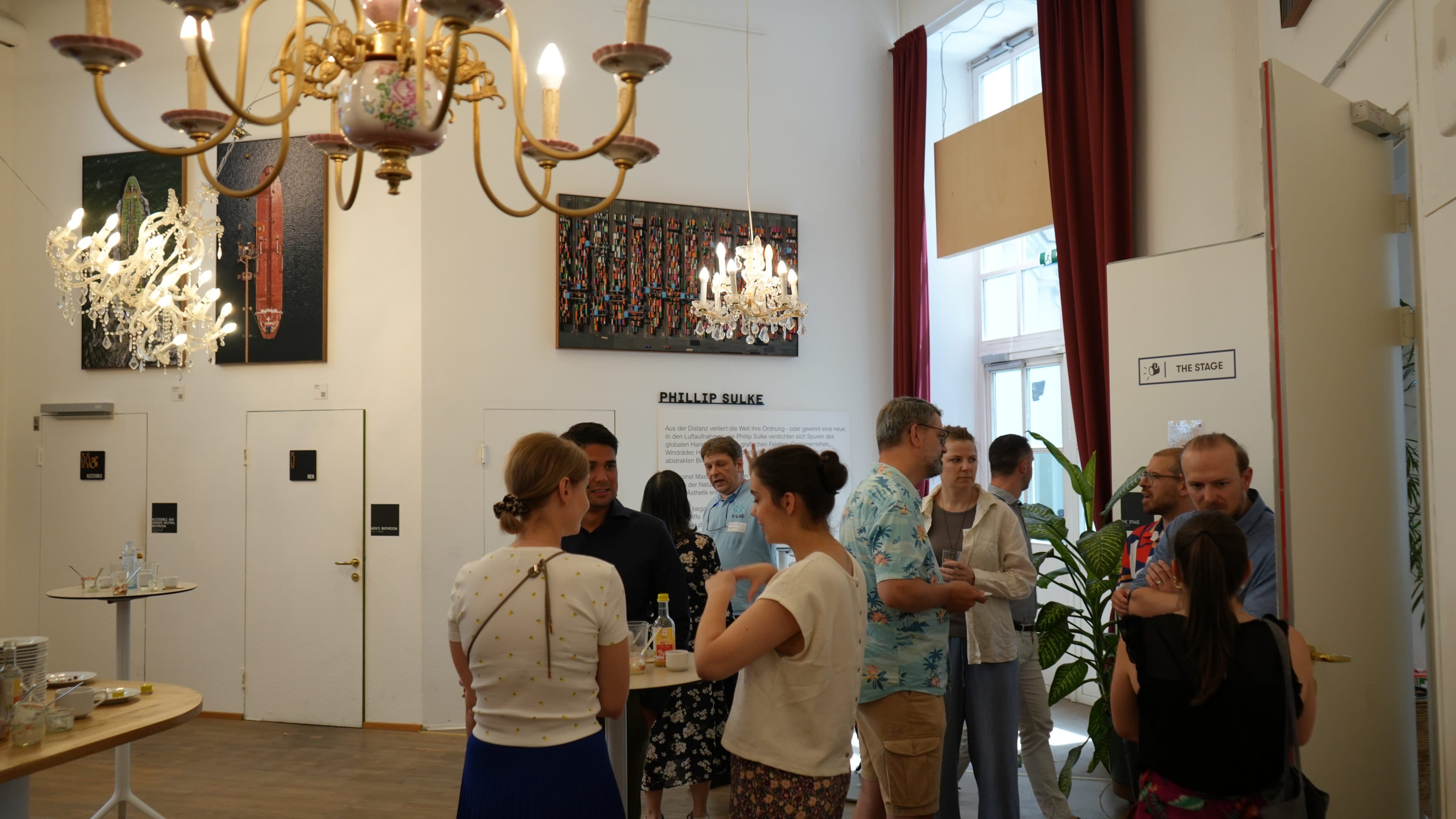Eine Gruppe von Menschen unterhält sich in einem hellen Raum mit Kronleuchtern, Kunstwerken an den Wänden und großen Fenstern. Einige stehen mit Getränken um Tische herum, was auf eine ungezwungene Veranstaltung oder ein Treffen hindeutet.
