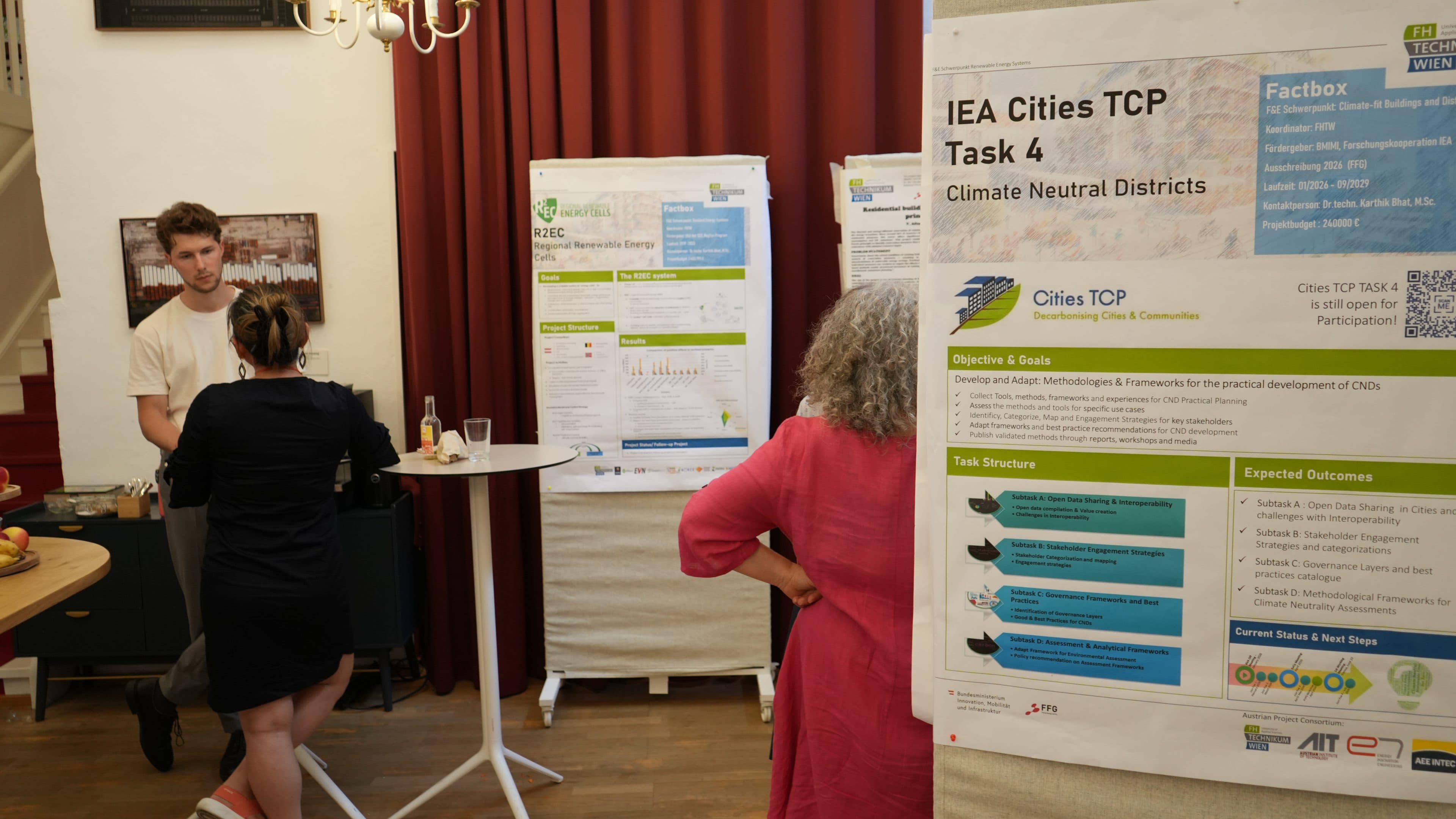 Eine Frau in einem rosa Kleid steht bei einer Veranstaltung mit Informationsständen vor einem Plakat mit der Aufschrift „IEA Cities TCP Task 4: Klimaneutrale Stadtteile“. Im Hintergrund unterhalten sich zwei Personen an einem Tisch.
