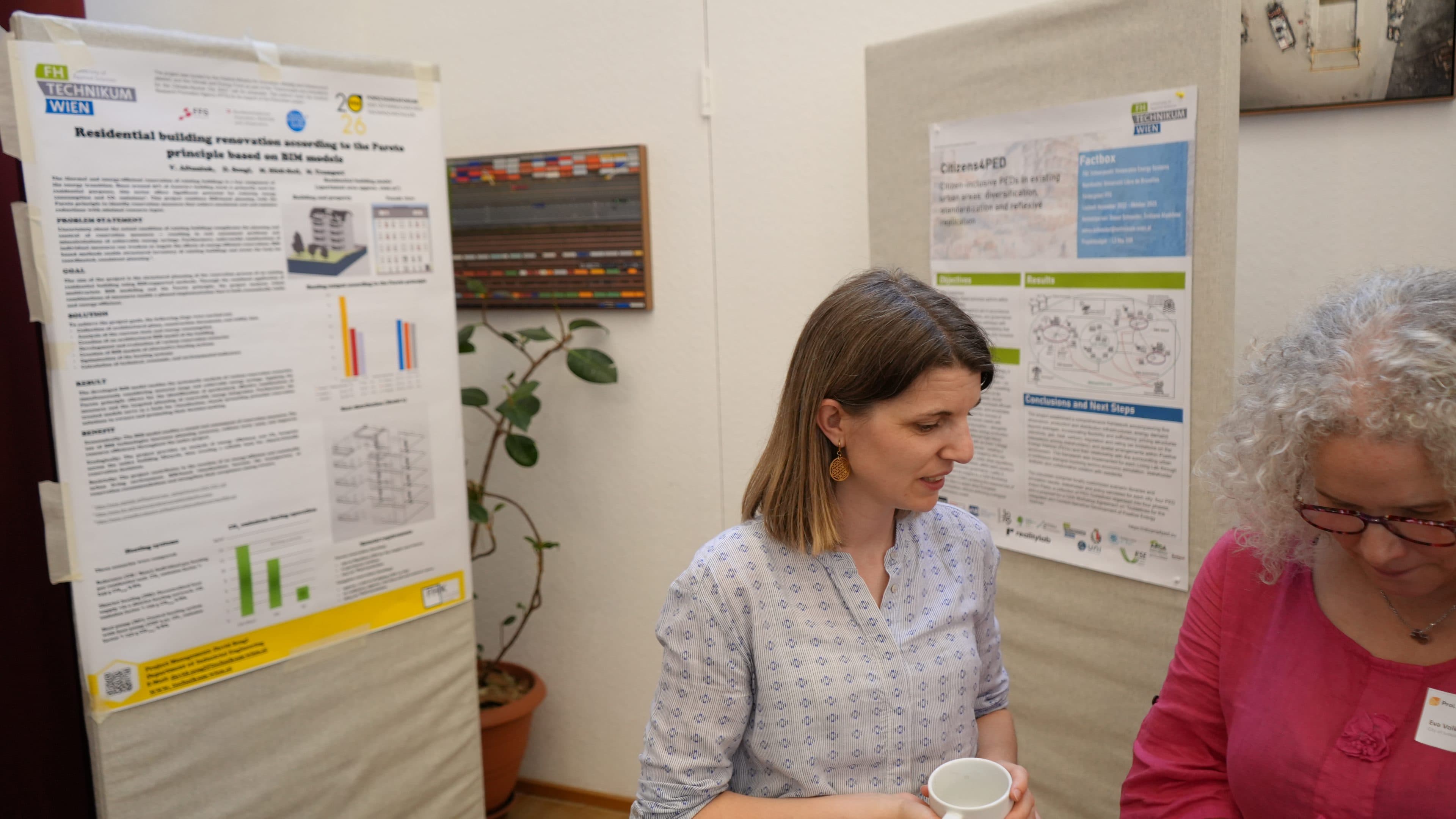 Zwei Frauen stehen vor Forschungsplakaten, die an Trennwänden im Rahmen einer Konferenz oder einer wissenschaftlichen Veranstaltung angebracht sind, und unterhalten sich. Eine der Frauen hält einen weißen Becher in der Hand; im Hintergrund sind eine Topfpflanze und eine farbige Wandverkleidung zu sehen.