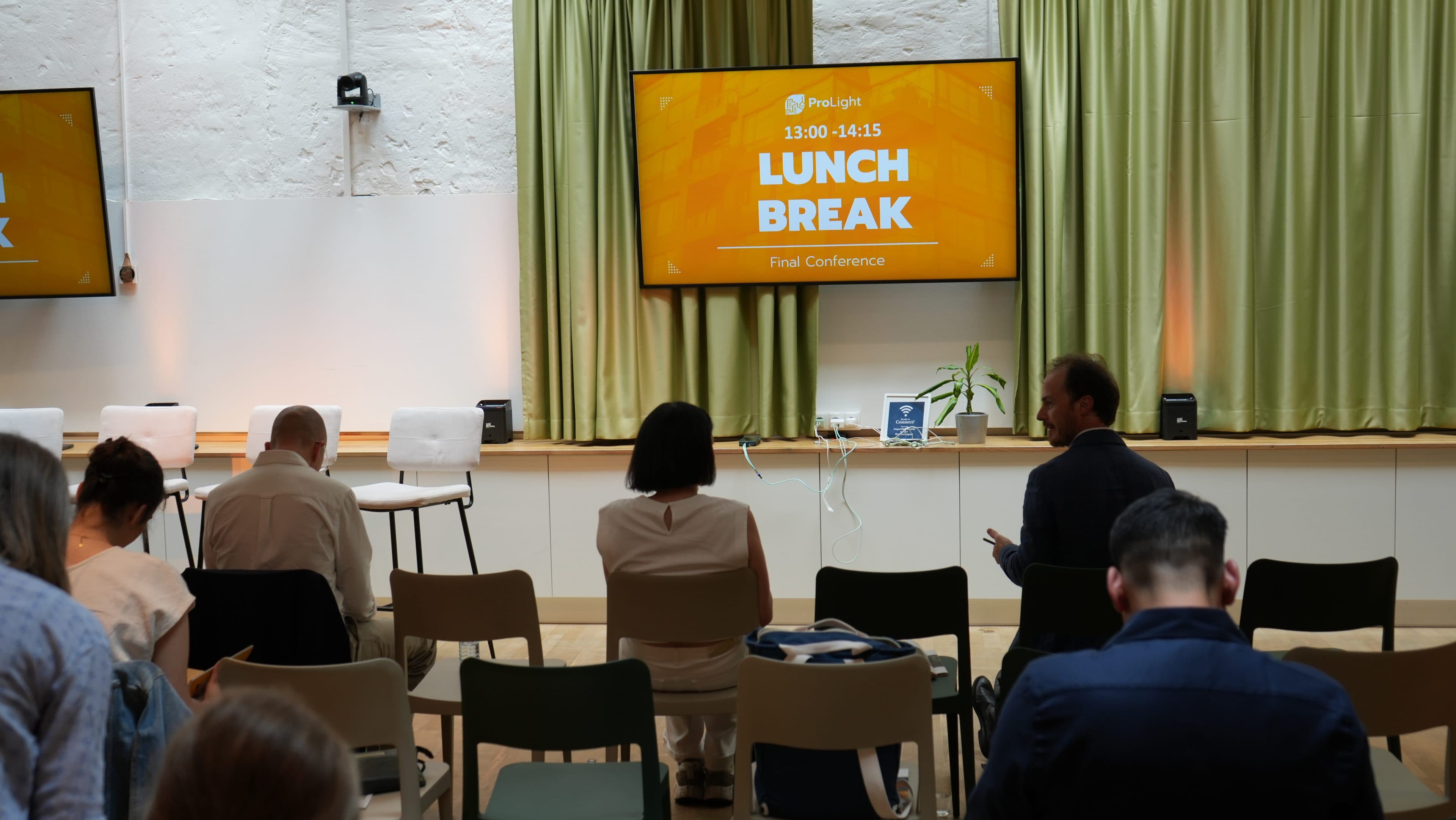 Ein Konferenzraum, in dem mehrere Personen sitzen und auf eine Leinwand blicken, auf der „Mittagspause“ sowie Details zur Konferenz zu sehen sind. Einige Stühle sind leer, und im Hintergrund sind die Fenster mit grünen Vorhängen verdeckt.