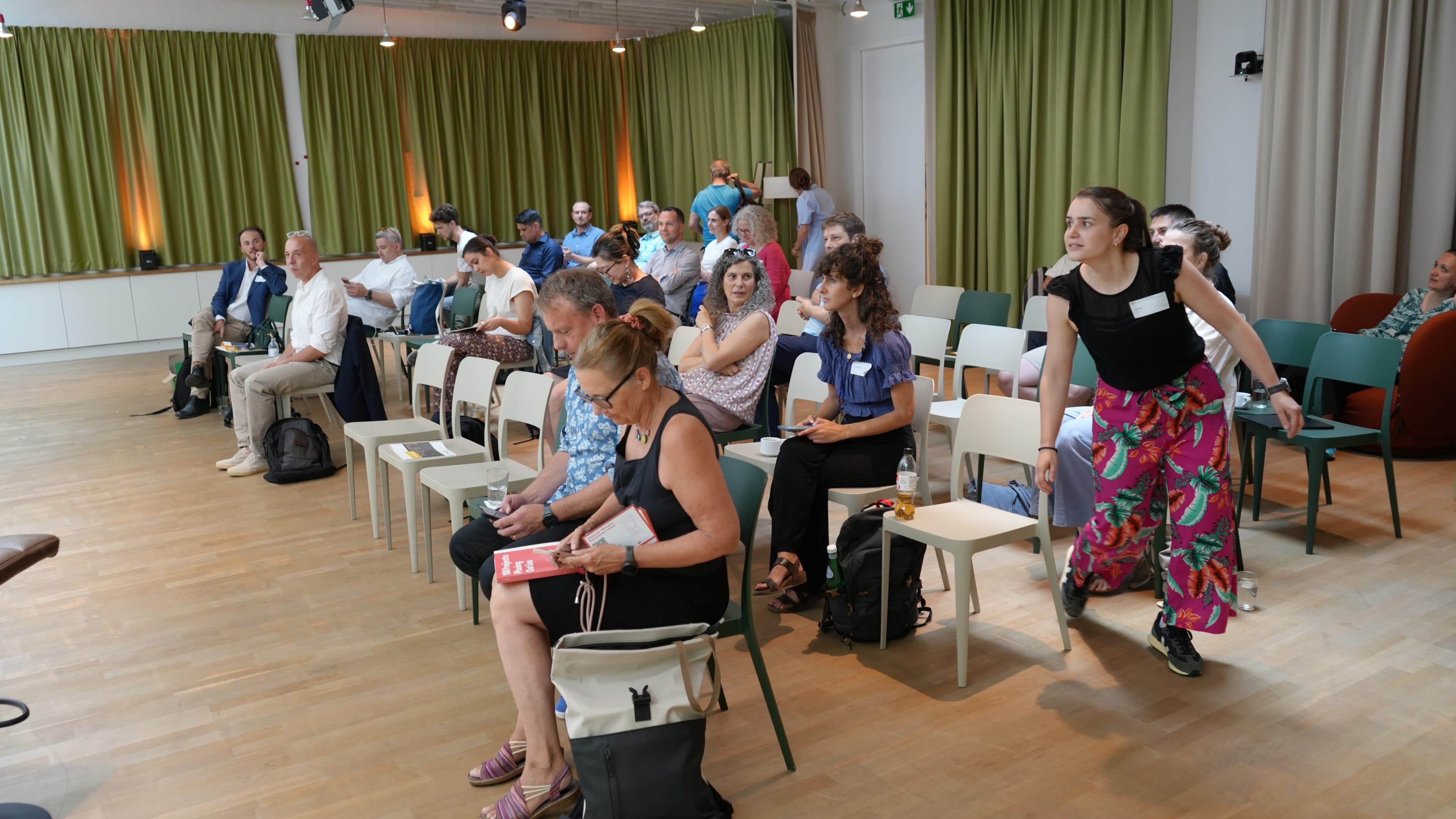 Eine Gruppe von Menschen sitzt in einem hellen, modernen Raum mit grünen Vorhängen und Holzböden und unterhält sich. Einige sitzen, andere stehen oder bewegen sich im Raum, und eine junge Frau in bunten Hosen geht zwischen den Stühlen hindurch.