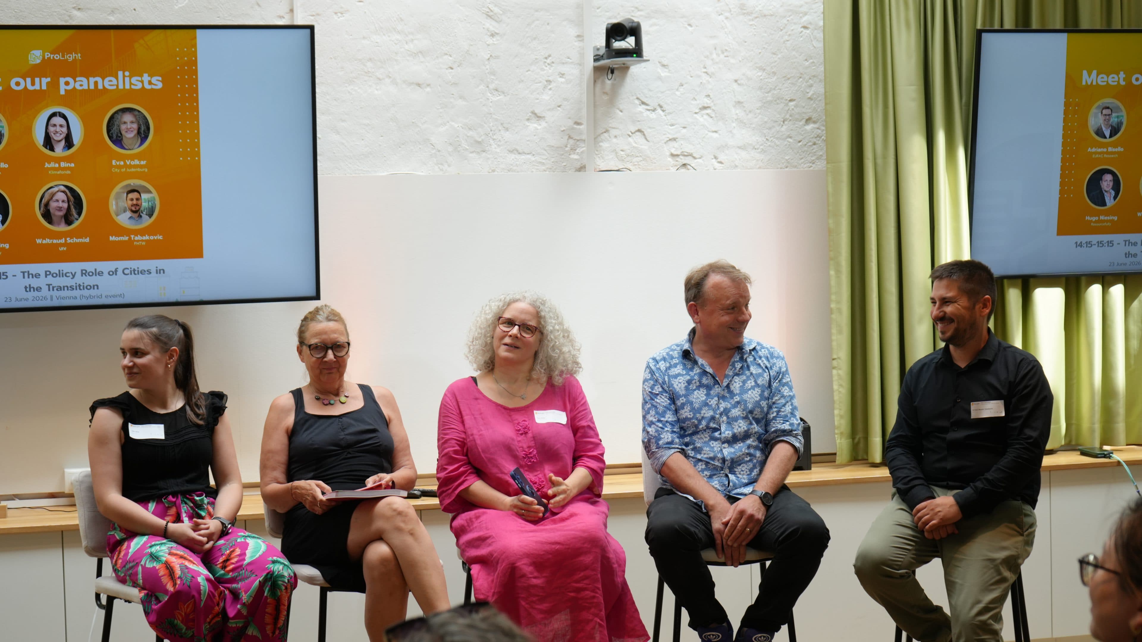 Fünf Diskussionsteilnehmer sitzen auf Hockern vor dem Publikum, im Hintergrund ist eine Leinwand zu sehen, auf der Veranstaltungsdetails und die Namen der Diskussionsteilnehmer angezeigt werden. Sie wirken aufmerksam und hören zu; sie sind leger bis halbformell gekleidet.