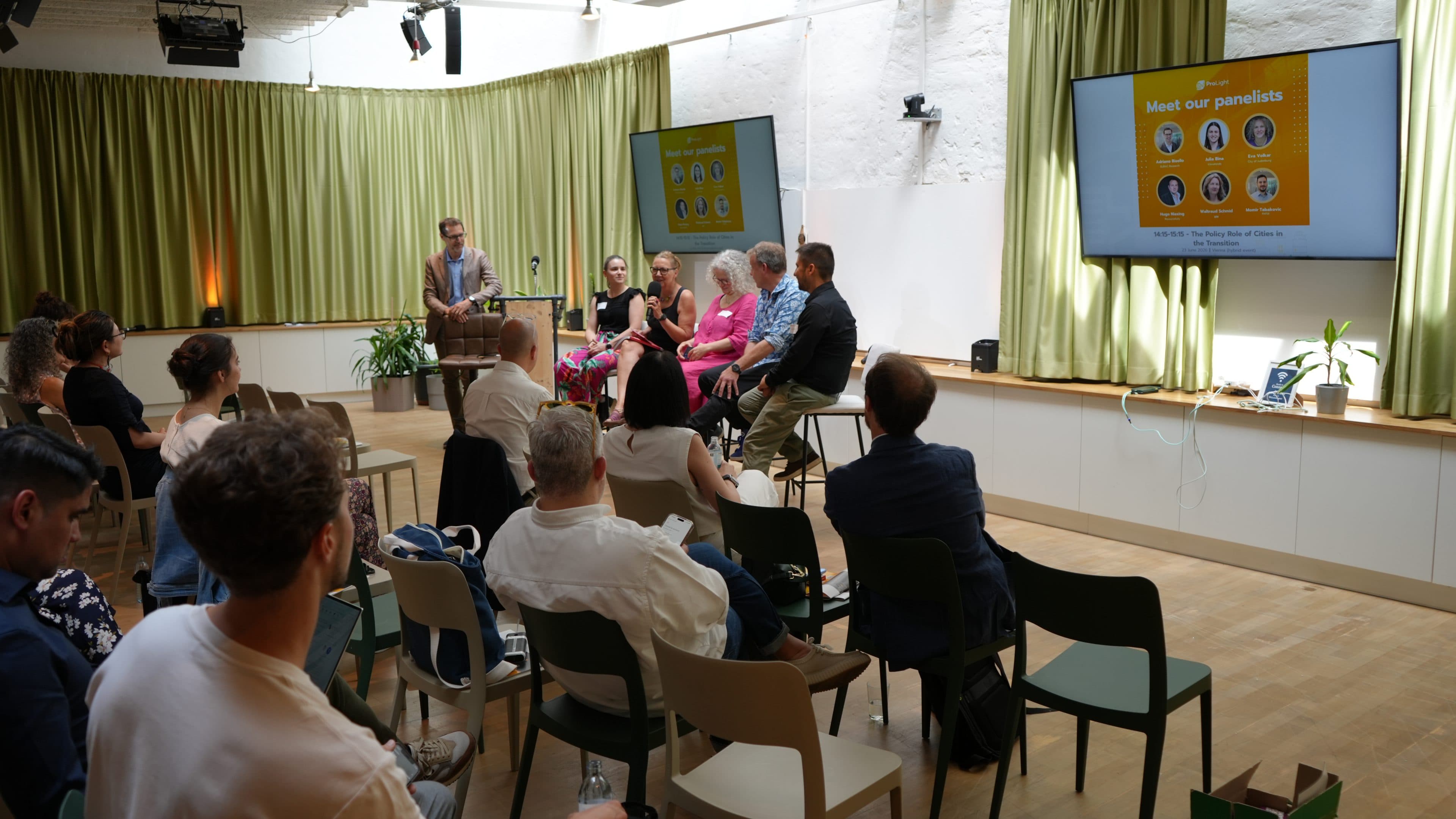 Auf der Bühne sitzt eine fünfköpfige Diskussionsrunde und spricht zu den Zuschauern in einem hellen Raum mit grünen Vorhängen. Auf zwei großen Bildschirmen werden Informationen zu den Diskussionsteilnehmern angezeigt. Das Publikum hört aufmerksam zu, wobei einige Stühle unbesetzt sind.