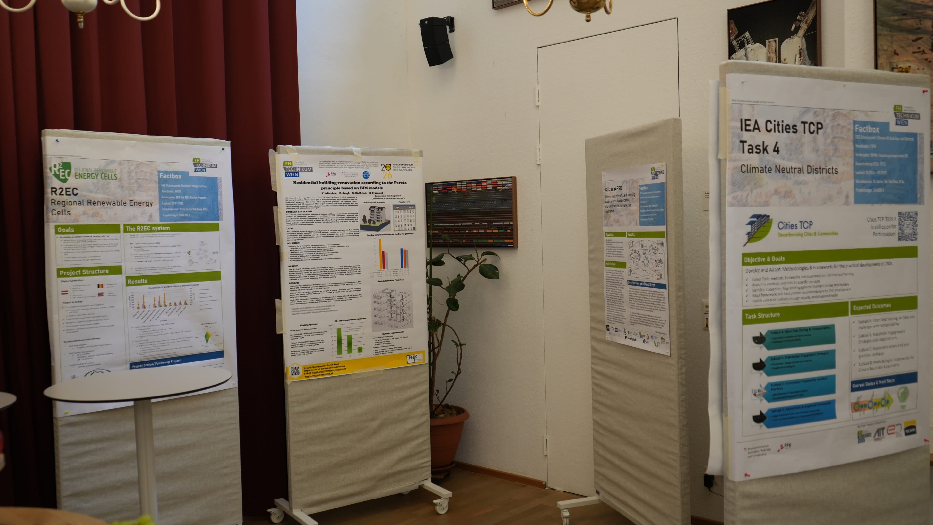 Vier Forschungsplakate zum Thema klimaneutrale Energie und Städte sind auf freistehenden Tafeln in einem Raum ausgestellt, der mit roten Vorhängen, einer Topfpflanze und einer weißen Wand mit einem an der Wand montierten Lautsprecher sowie einigen Wanddekorationen ausgestattet ist.