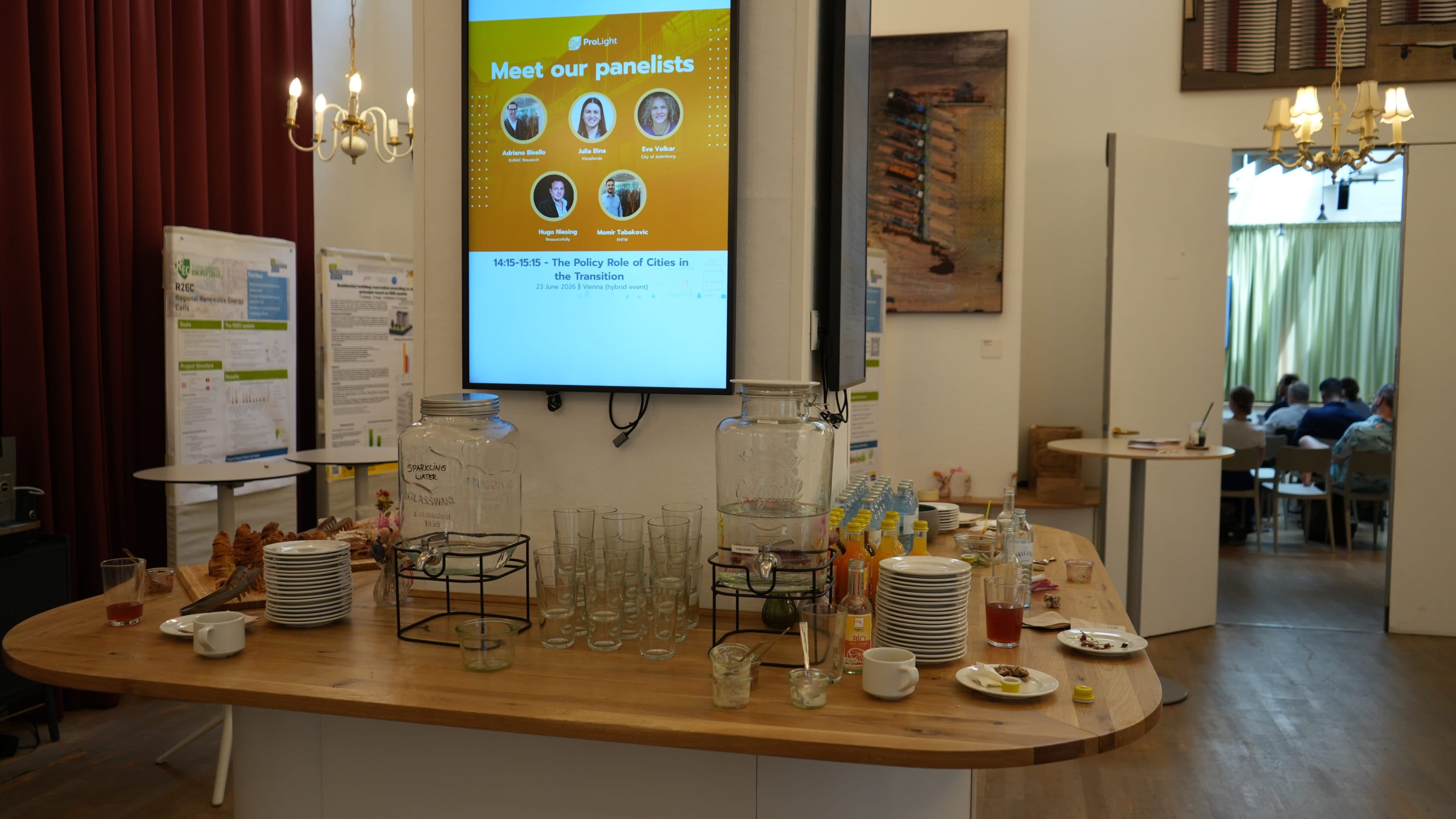 Auf einem Holztisch stehen leere Gläser, Teller, Krüge, Snacks und Getränke. Dahinter ist auf einem digitalen Bildschirm die Aufschrift „Lernen Sie unsere Podiumsteilnehmer kennen“ zu sehen, zusammen mit Fotos und Informationen zu einer Podiumsdiskussion. Im Hintergrund haben sich Menschen in einem Raum versammelt.
