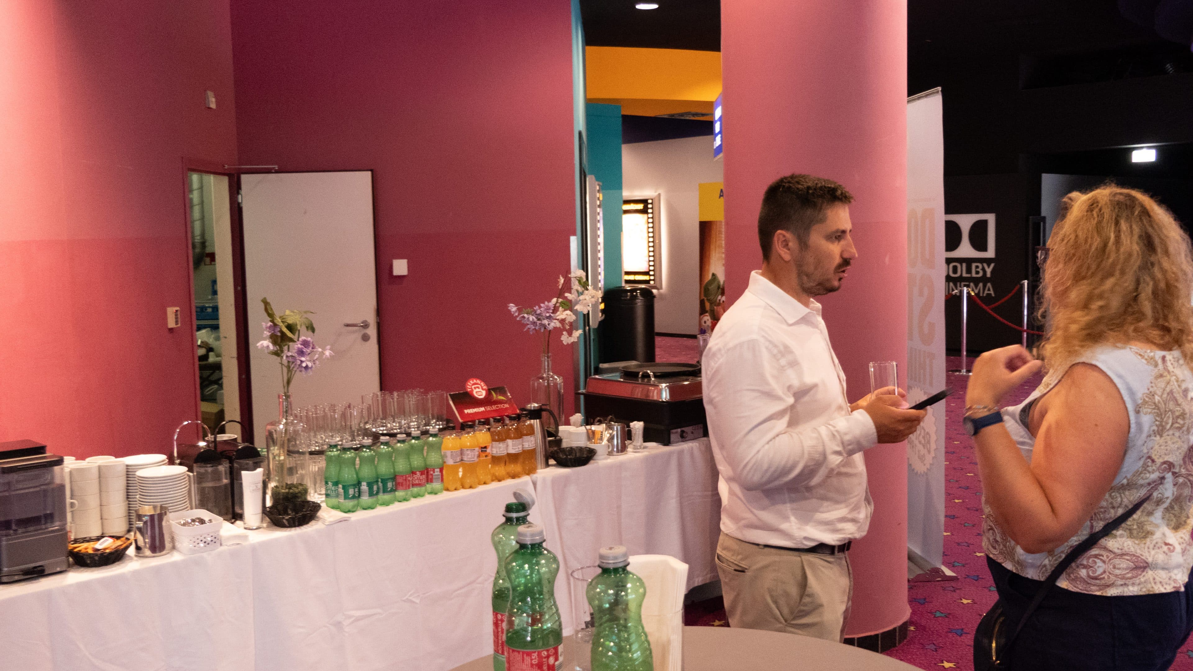 Zwei Personen unterhalten sich in einem farbenfrohen Raum mit rosa Säulen und einem gemusterten Teppich in der Nähe eines Buffets mit Getränken, Bechern und Snacks.