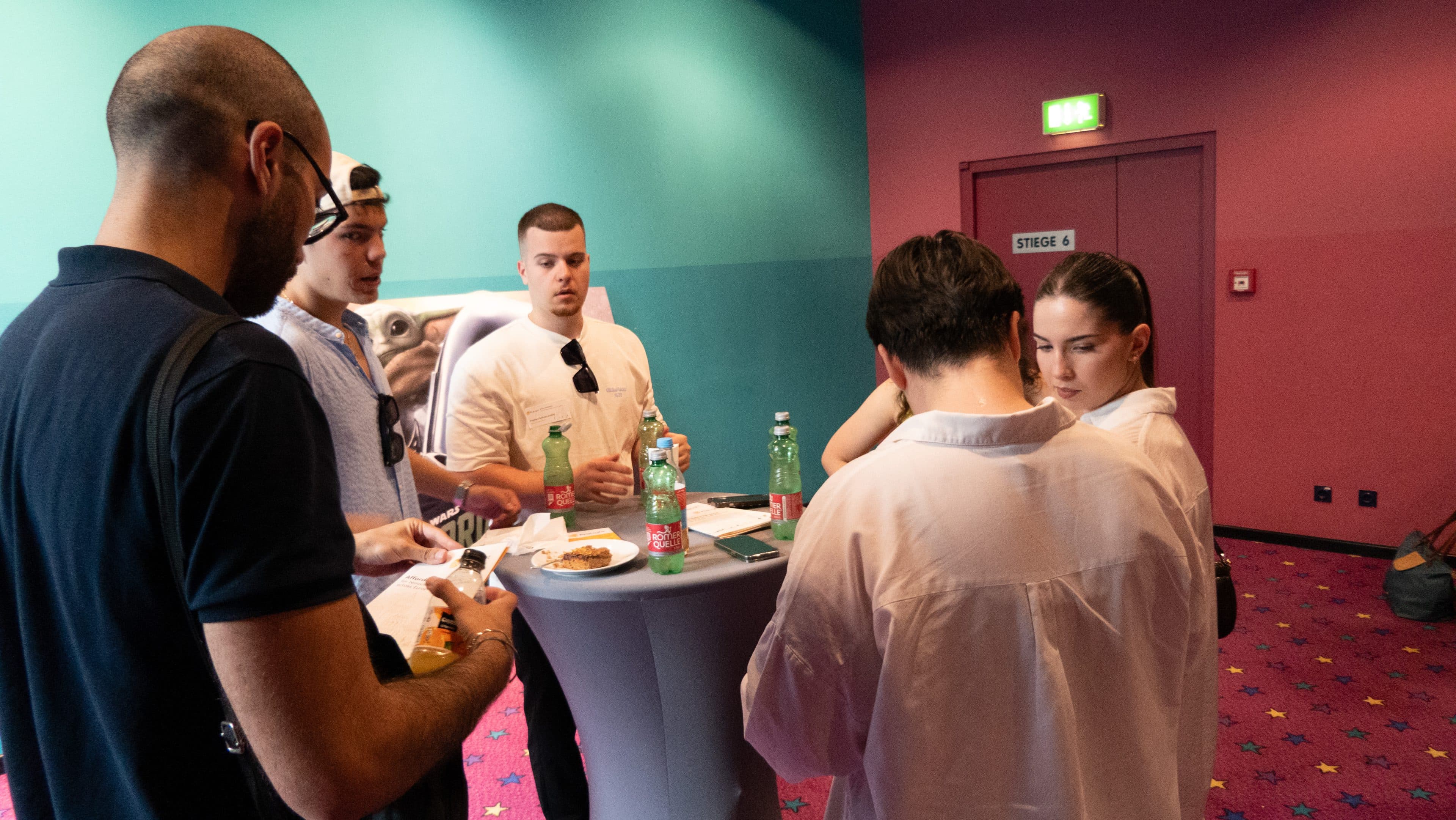 Fünf Personen stehen um einen hohen Tisch herum, auf dem Getränke und Snacks stehen, in einem hell beleuchteten Raum mit violetten Wänden und einem gemusterten Teppich. Einige unterhalten sich, während andere auf ihre Handys oder ihr Essen schauen.