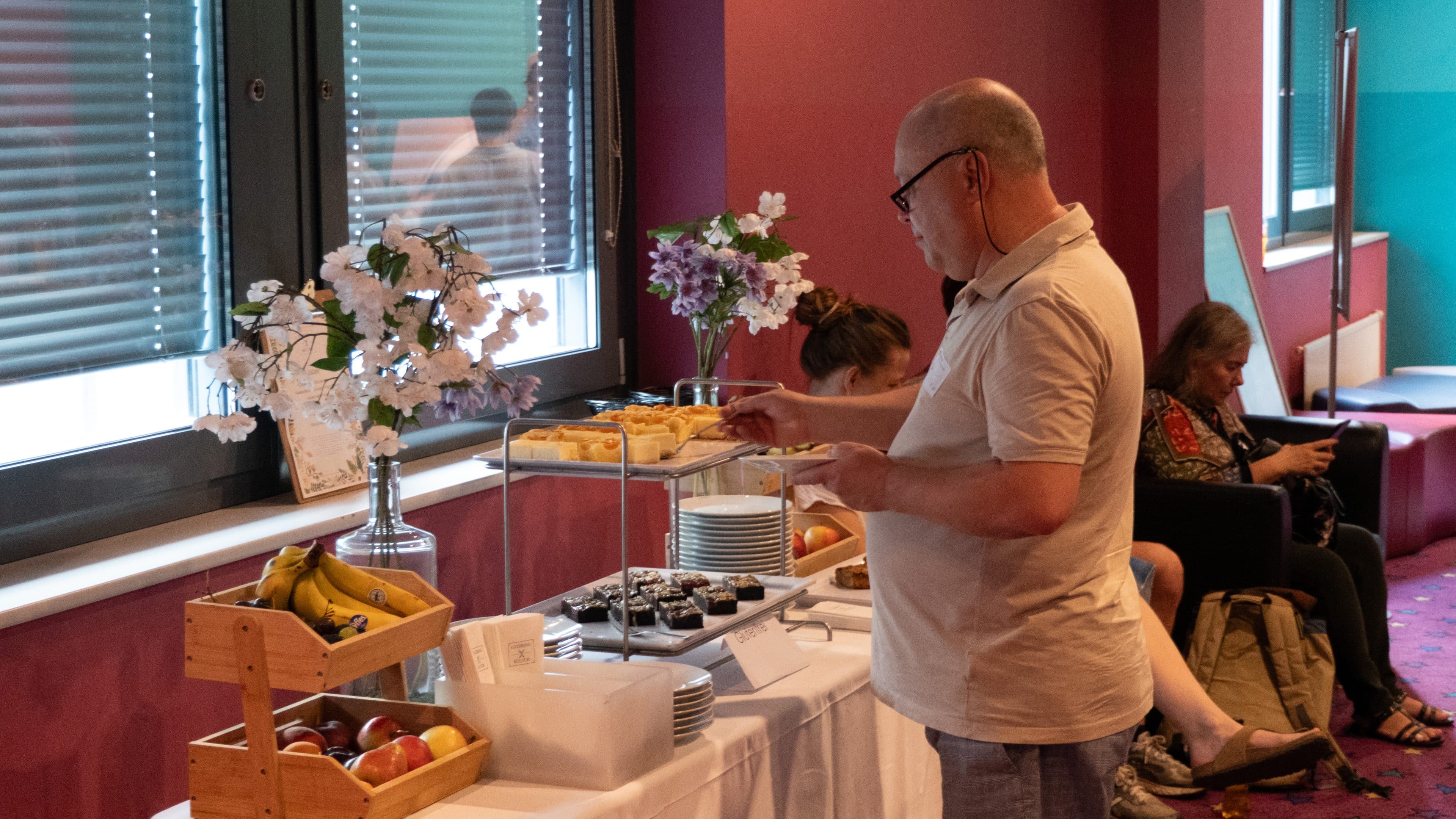 Ein Mann bedient sich am Buffettisch mit Gebäck, Obst und Desserts, während im Hintergrund andere sitzen. Der Raum hat burgunderrote Wände und ist mit Vasen mit weißen und violetten Blumen geschmückt.