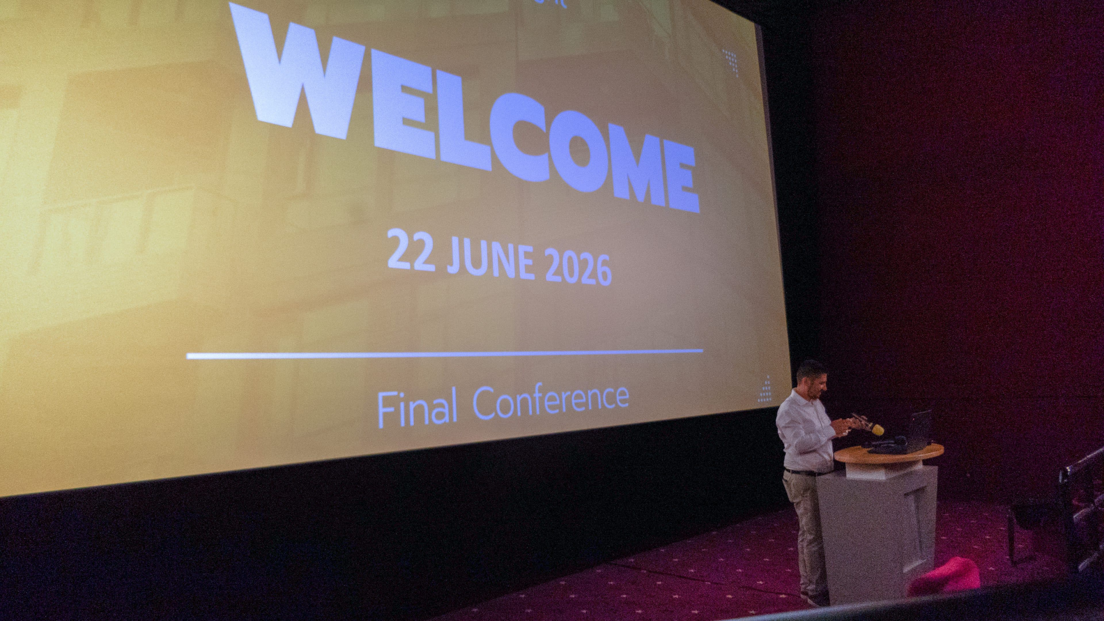 Ein Redner steht auf einem Podium in einem dunklen Saal neben einer großen Leinwand, auf der in fetten Buchstaben „WELCOME, 22. Juni 2026, Abschlusskonferenz“ zu lesen ist.