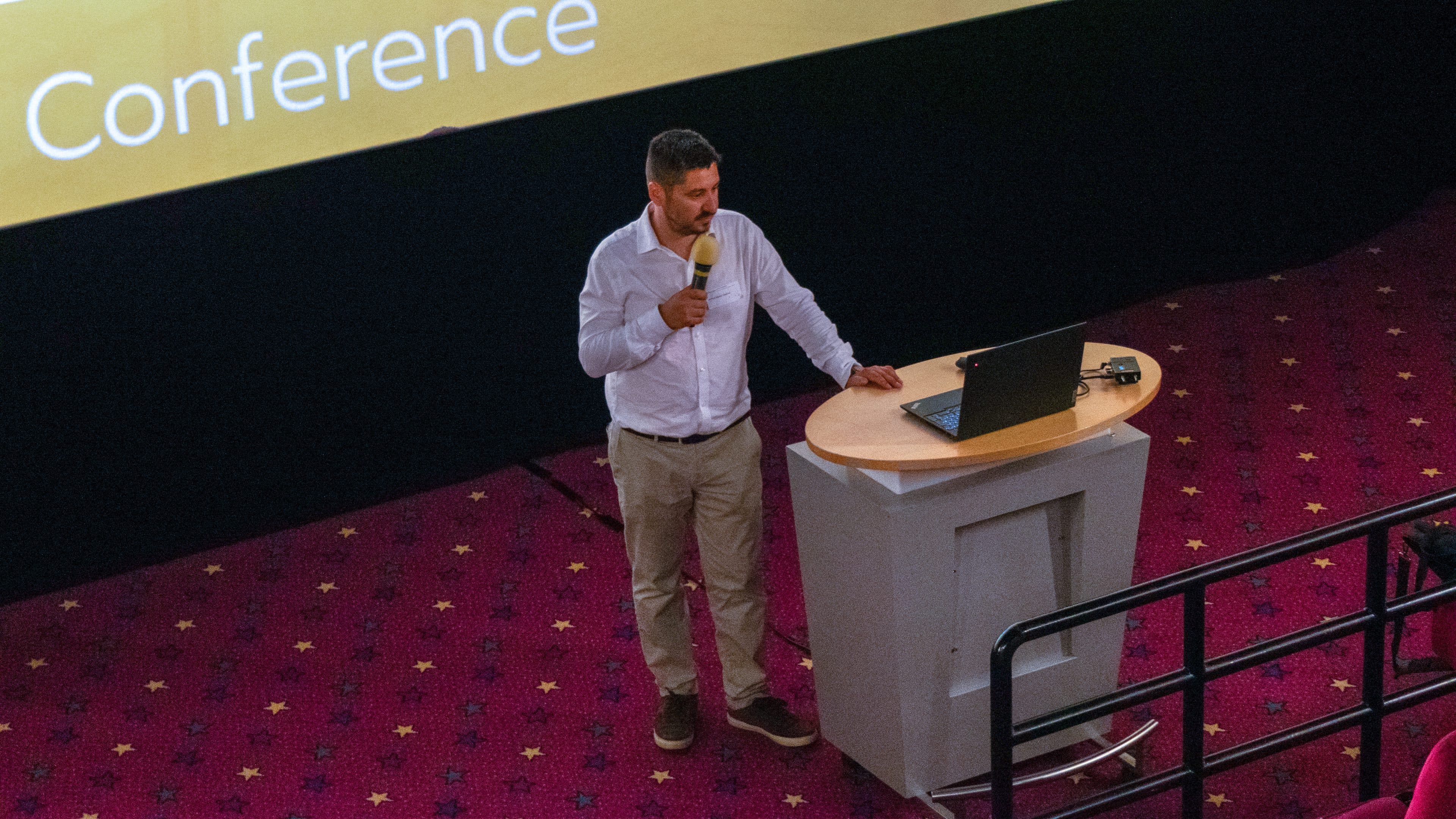 Ein Mann in einem weißen Hemd steht mit einem Laptop am Rednerpult, hält ein Mikrofon in der Hand und hält auf einer Konferenz eine Rede. Auf der Bühne liegt ein roter Teppich mit Sternenmuster, und im Hintergrund ist eine große Leinwand zu sehen.
