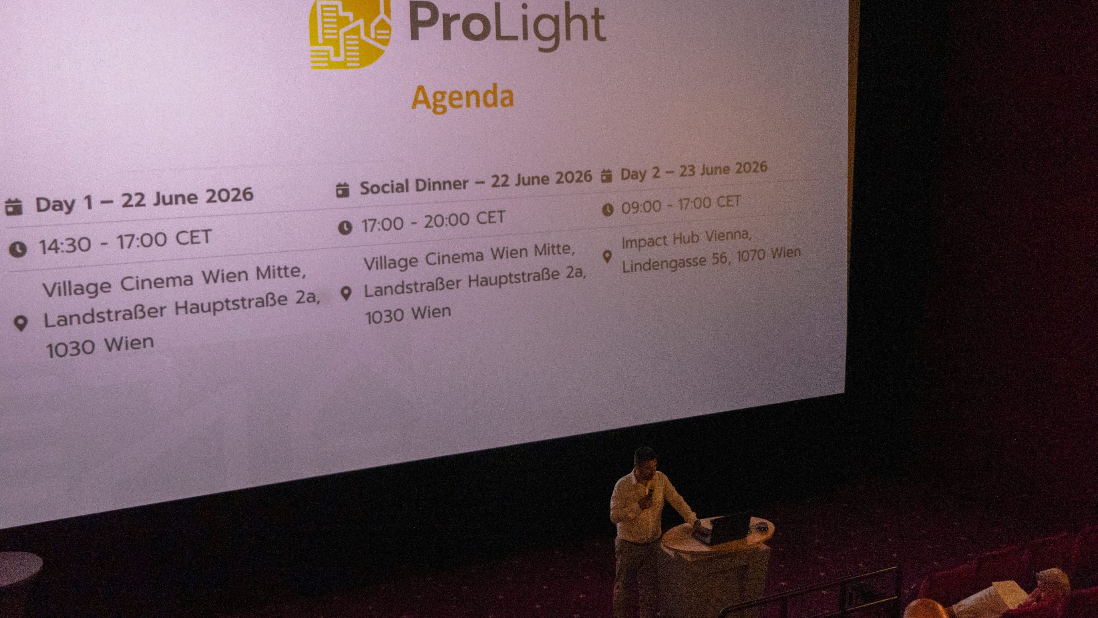 Ein Redner steht an einem Rednerpult in einem abgedunkelten Saal und spricht zu den Zuhörern. Auf der großen Leinwand hinter ihm wird das ProLight-Programm mit den Terminen, Uhrzeiten, Veranstaltungsorten und Einzelheiten der Veranstaltungen in Wien im Juni 2026 angezeigt.