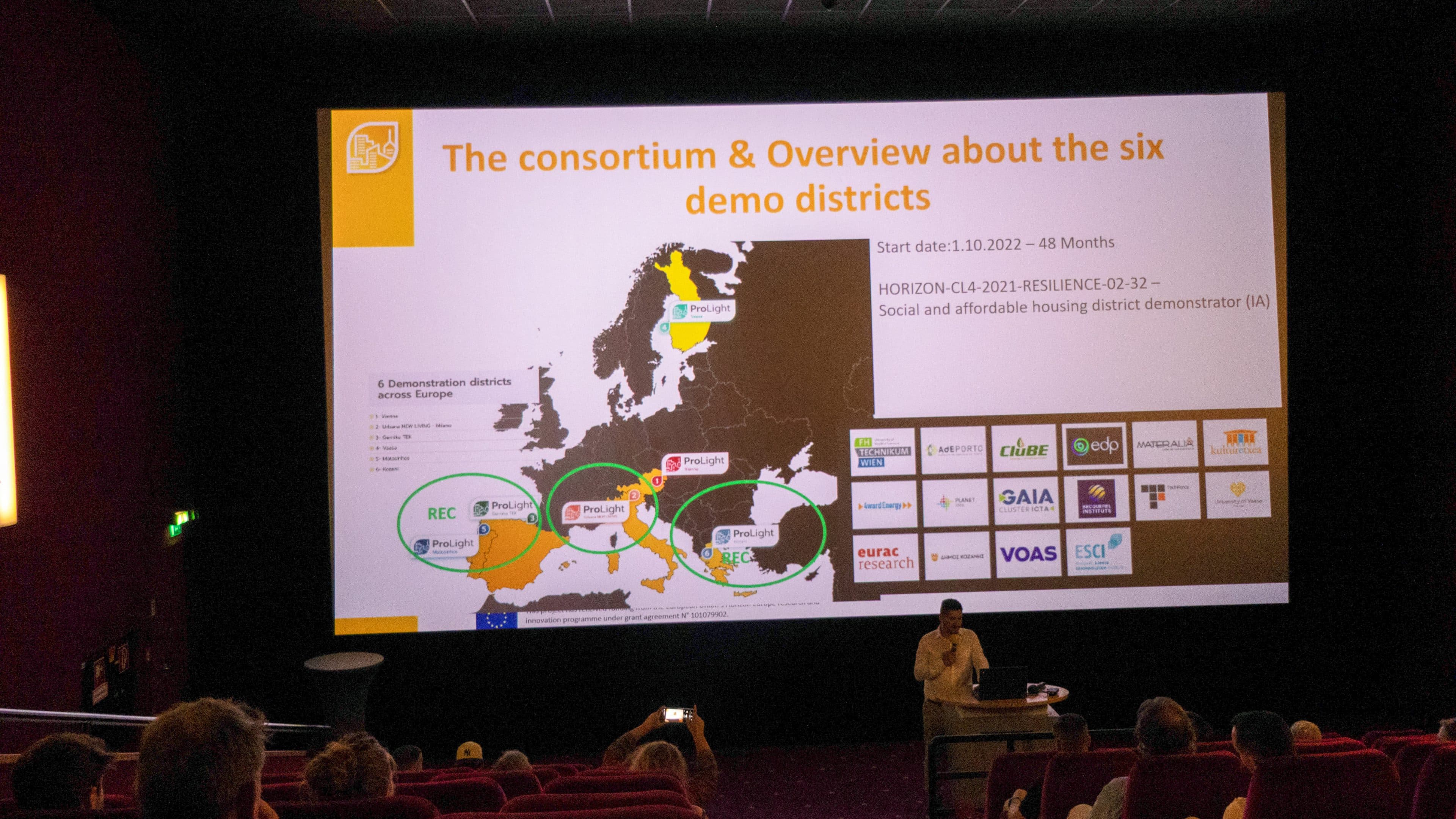 Ein Referent steht vor einer großen Leinwand in einem abgedunkelten Saal und zeigt im Rahmen einer Übersicht über ein Forschungskonsortium eine Karte von Europa, Logos und Text zu sechs Modellwohnvierteln.