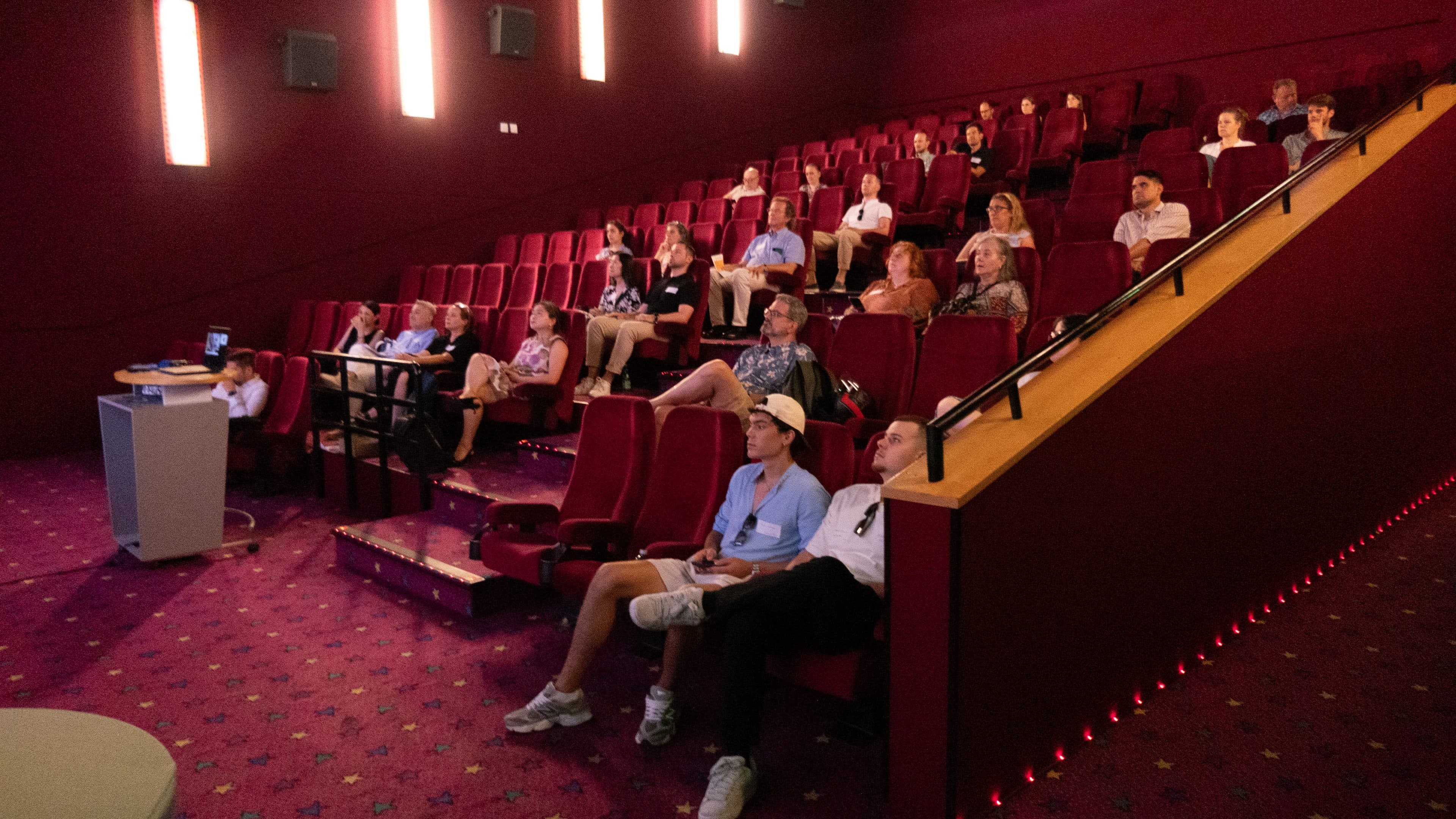 Eine Gruppe von Menschen sitzt in einem schwach beleuchteten Kino mit roten Sitzen und Teppichboden und blickt aufmerksam auf eine Leinwand, die nicht zu sehen ist. Die meisten Plätze sind besetzt, und das Publikum wirkt konzentriert.