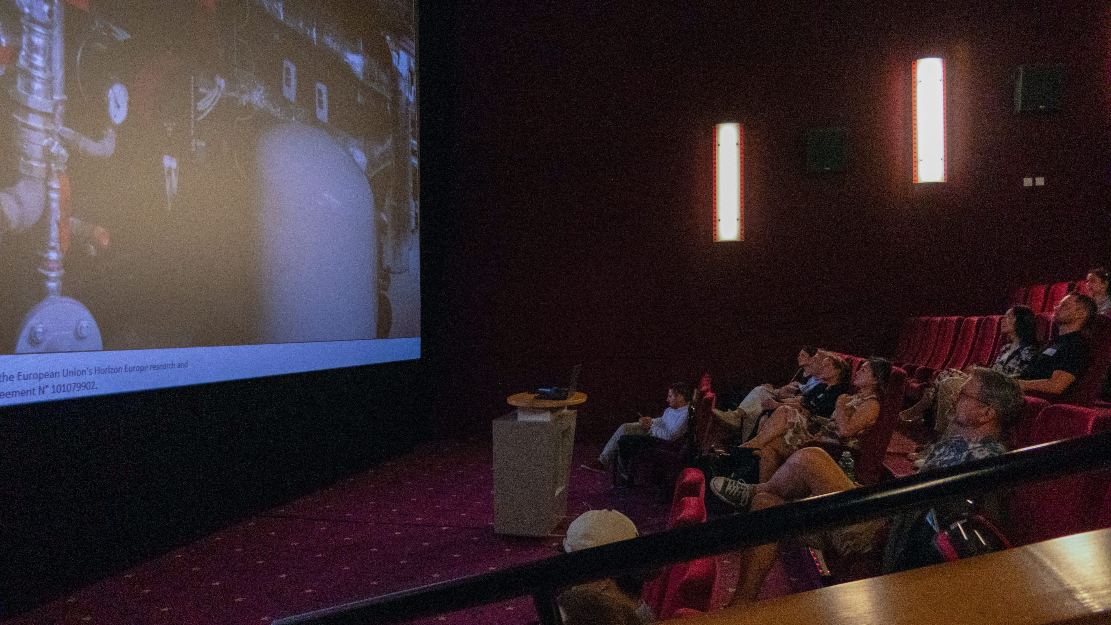 In einem schummrig beleuchteten Kino sitzen Menschen auf roten Kinosesseln und blicken auf eine große Leinwand. Auf der Leinwand ist eine Industrieszene zu sehen, und der Raum verfügt über Wände mit vertikalen Leuchten sowie ein kleines Podium in der Nähe der Leinwand.