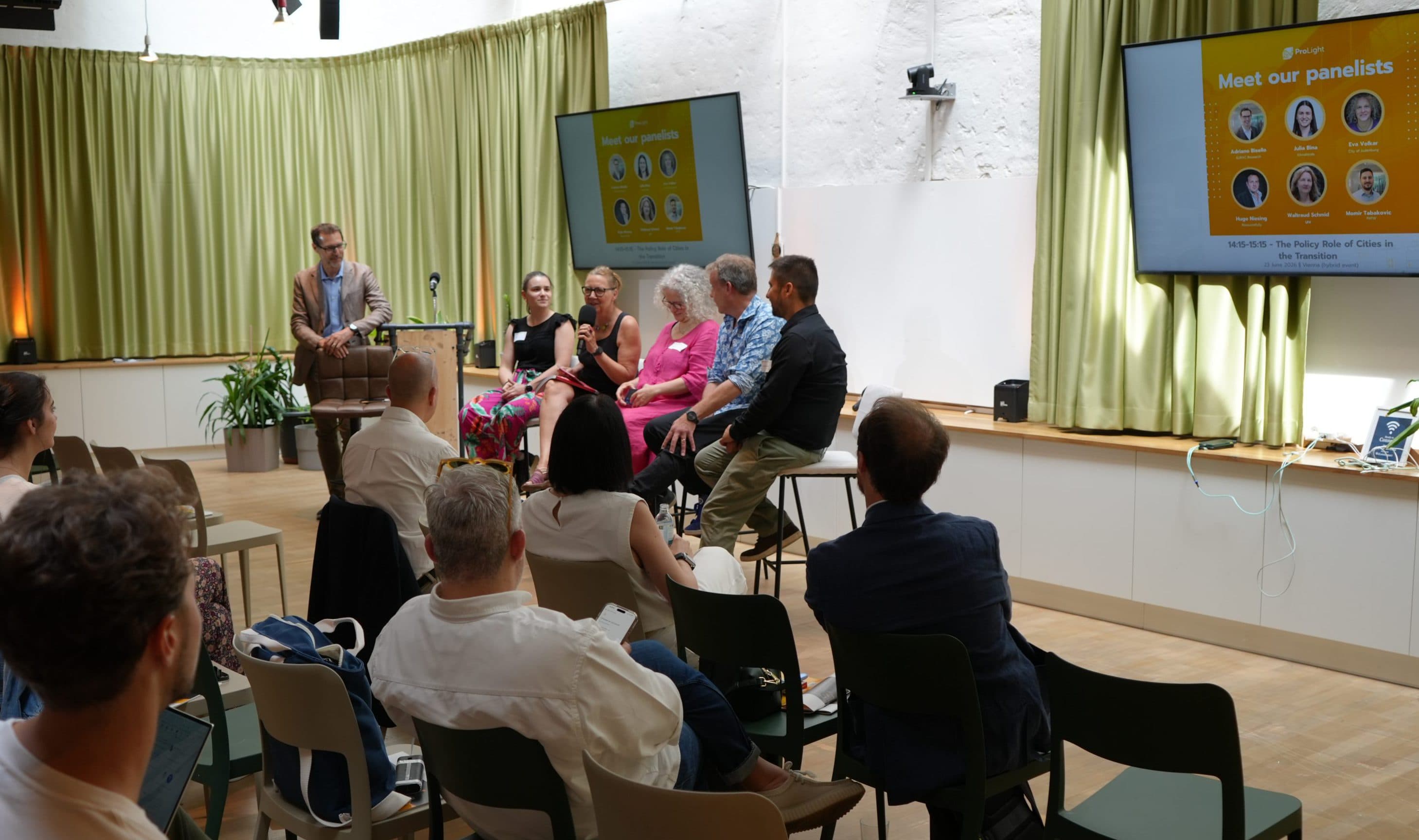 Eine fünfköpfige Diskussionsrunde spricht auf der Bühne vor einem Publikum in einem hellen Raum mit grünen Vorhängen. Auf zwei Bildschirmen werden Informationen zu den Diskussionsteilnehmern angezeigt. Das Publikum sitzt in Reihen und hört aufmerksam zu.