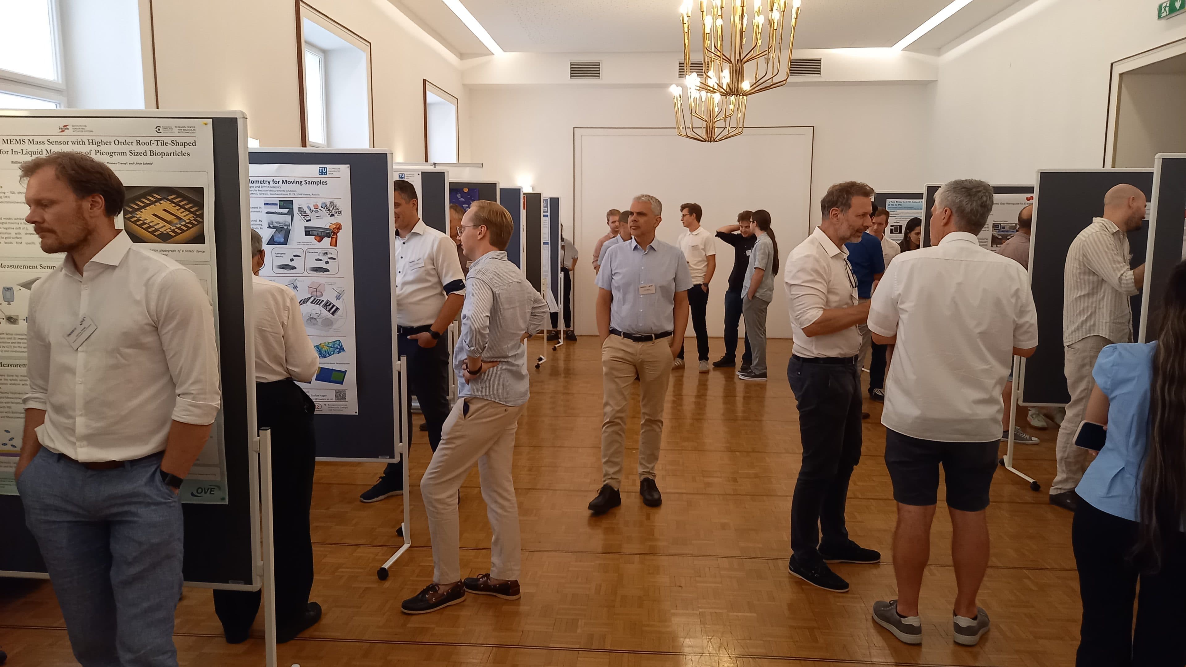 Eine Gruppe von Menschen in einem gut beleuchteten Raum diskutiert und betrachtet während einer Konferenz oder einer wissenschaftlichen Veranstaltung wissenschaftliche Poster, die an Tafeln ausgestellt sind. Die meisten Teilnehmer tragen Business-Casual-Kleidung.
