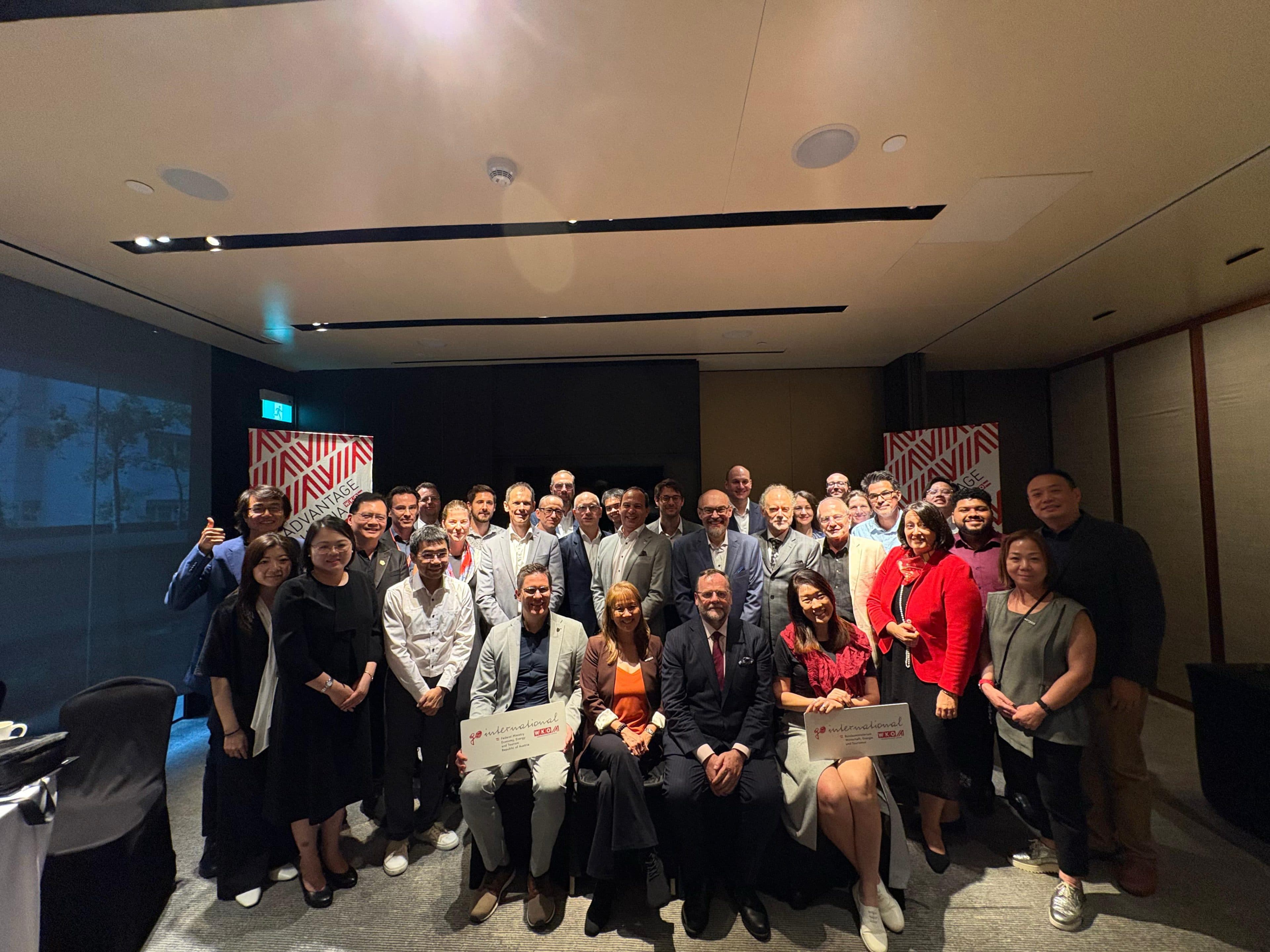 Eine große Gruppe von Menschen in Business-Kleidung posiert bei einer Veranstaltung gemeinsam in einem Raum für ein Gruppenfoto. Einige sitzen vorne, während andere hinter ihnen stehen, Schilder in den Händen halten und in die Kamera lächeln.