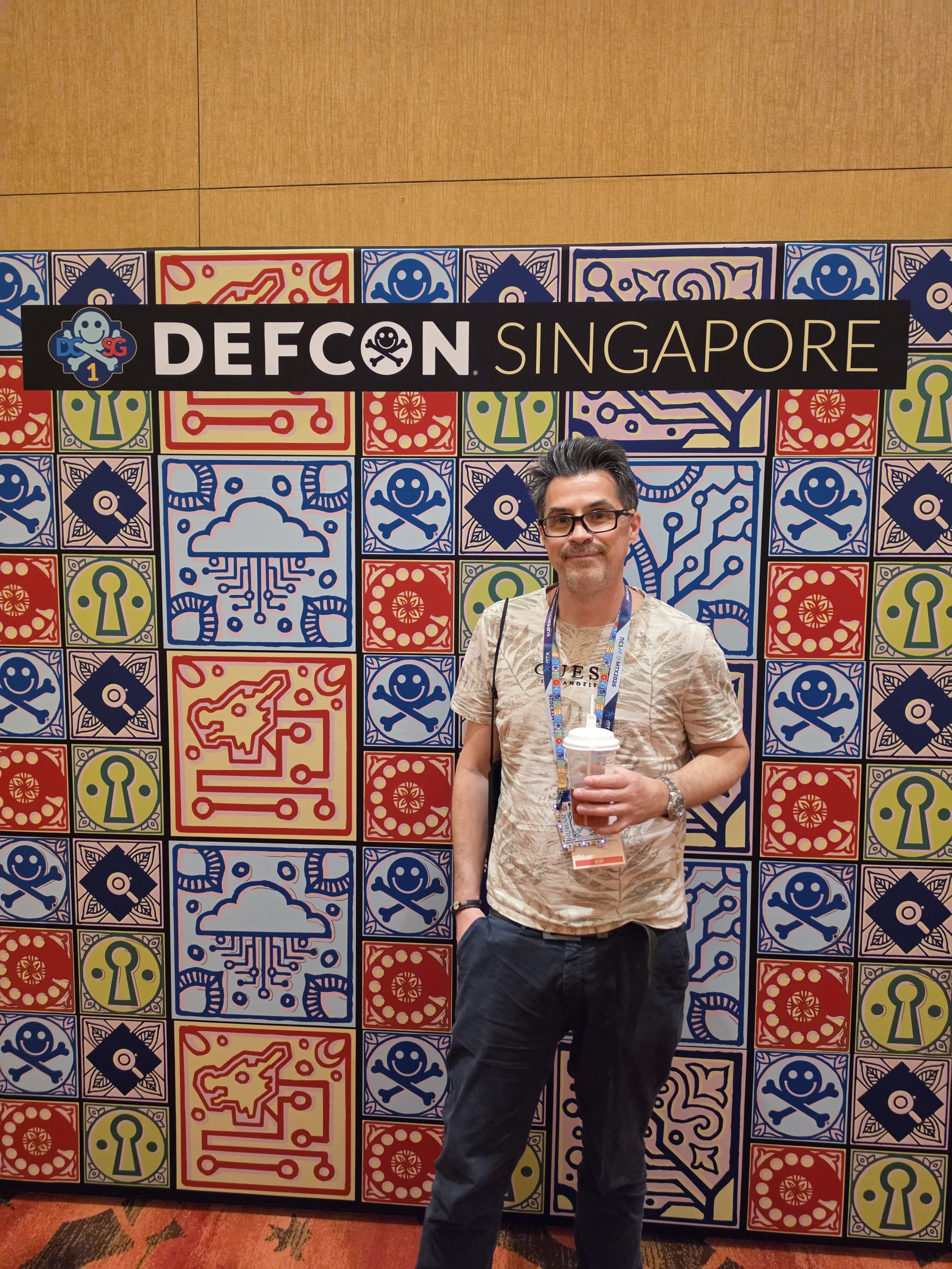 Ein Mann steht vor einer bunt gemusterten Wand, auf der „DEFCON SINGAPORE“ steht. Er hält ein Getränk in der Hand und trägt ein Schlüsselband. Die Wand ist mit Motiven aus den Bereichen Technik und Elektronik in den Farben Rot, Blau und Gelb verziert.