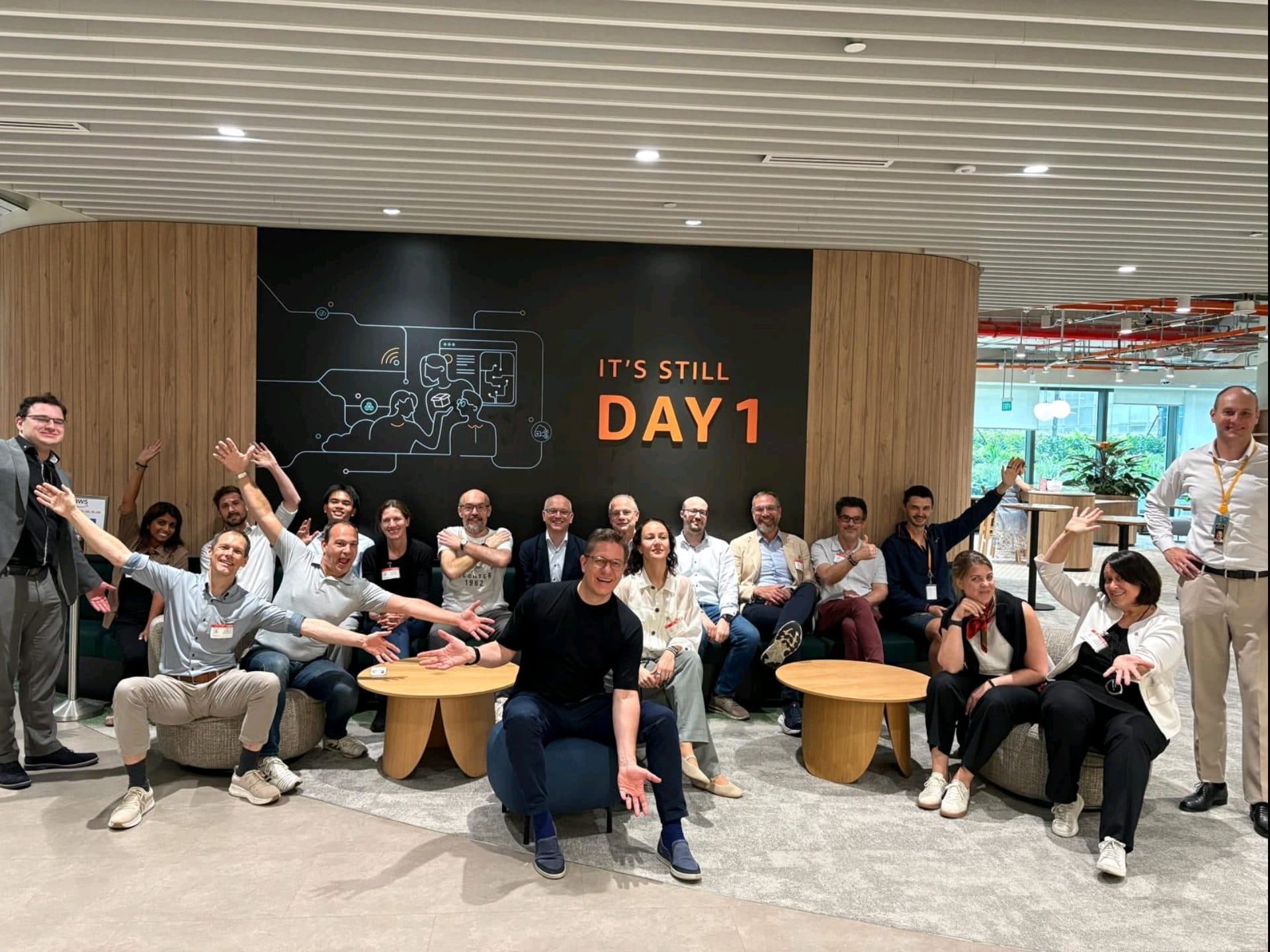 Eine Gruppe von etwa 20 Personen in Freizeitkleidung sitzt und steht um Holztische in einem Büroraum herum und lächelt und posiert begeistert vor einer Wand, auf der „ITS STILL DAY 1.“ steht.