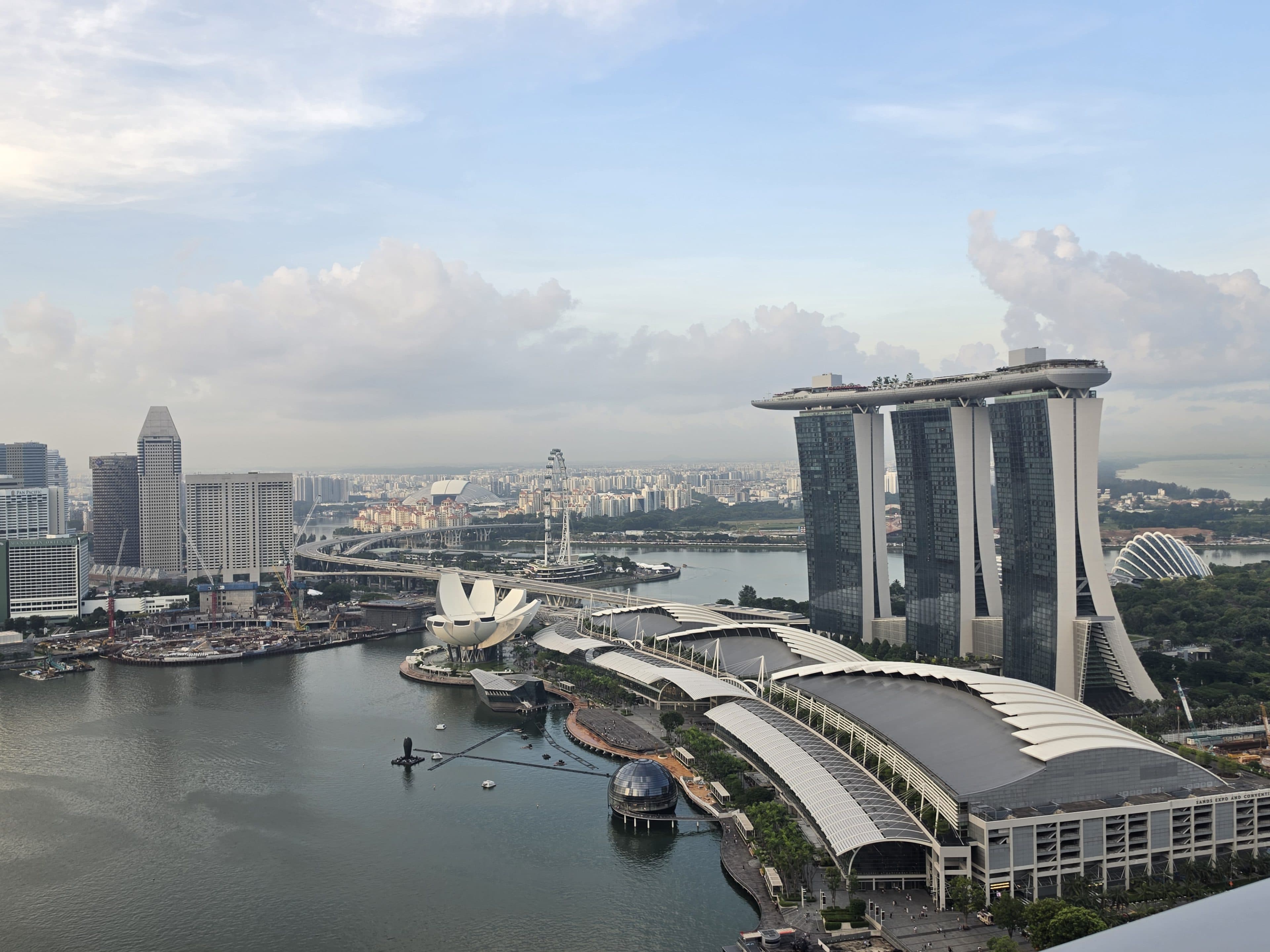 Ein Panoramablick auf Marina Bay in Singapur mit dem berühmten Marina Bay Sands Hotel, dem ArtScience Museum und der umliegenden Skyline unter einem teilweise bewölkten Himmel.