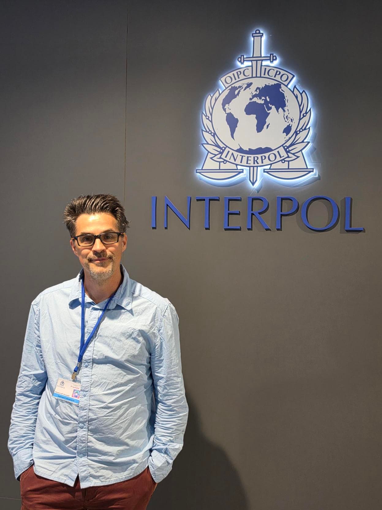 Ein Mann mit Brille, blauem Hemd und einem gut sichtbaren Ausweis steht vor einer Wand mit dem INTERPOL-Logo und -Emblem.
