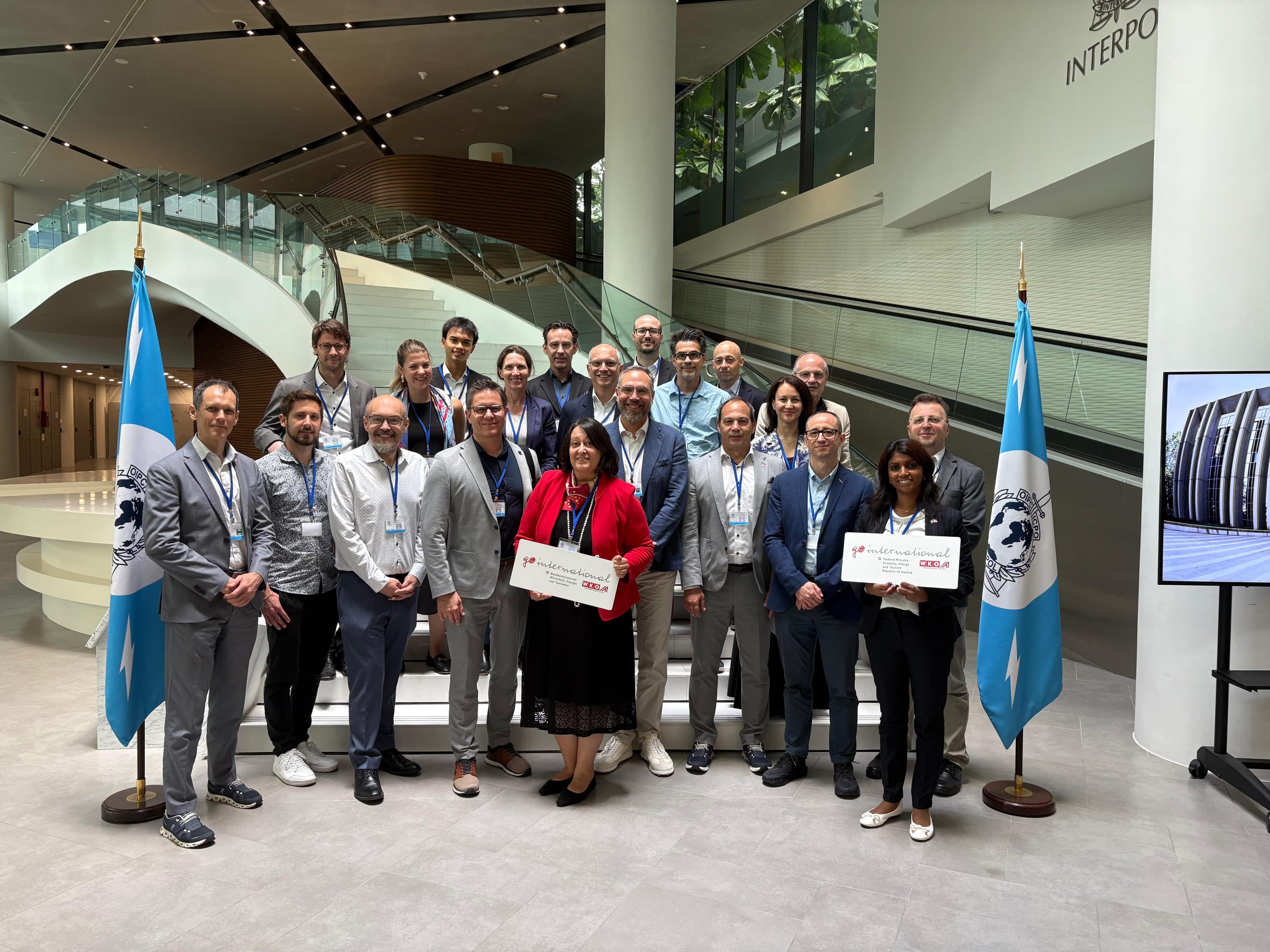 Eine Gruppe von Menschen posiert in einem Innenraum zwischen zwei INTERPOL-Flaggen für ein Foto. Einige halten Schilder mit der Aufschrift „CONGRATS“ in der Hand. Im Hintergrund sind eine moderne Treppe und eine Glaswand zu sehen.