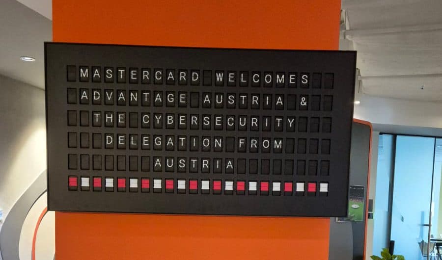 An einer orangefarbenen Wand hängt eine schwarze Anschlagtafel mit weißer Schrift, auf der zu lesen ist: „Mastercard heißt Advantage Austria und die Cybersicherheitsdelegation aus Österreich willkommen“. In der Nähe sind Topfpflanzen und Büromöbel zu sehen.