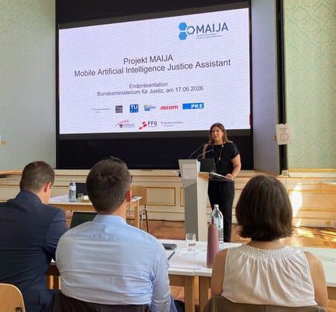 Eine Frau steht am Rednerpult und hält vor einem Publikum in einem Konferenzraum einen Vortrag zum Thema „Projekt MAIJA: Mobiler KI-Assistent für die Justiz“, während hinter ihr ein großer Bildschirm und Logos zu sehen sind.