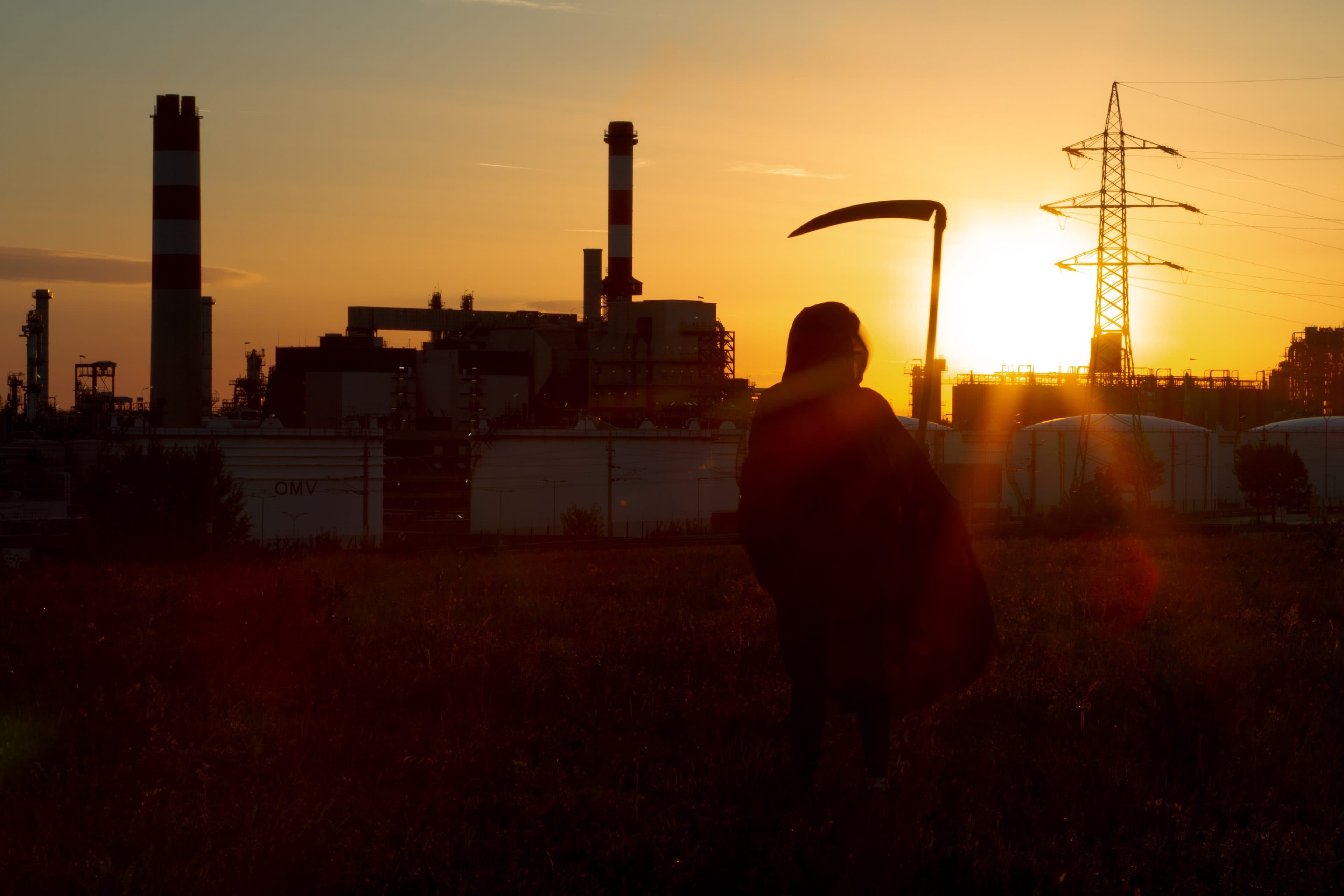 Vor einer Industrieanlage steht bei Sonnenuntergang die Silhouette einer Gestalt mit einer Sense in der Hand; vor dem orangefarbenen Himmel sind Schornsteine und Stromleitungen zu sehen.