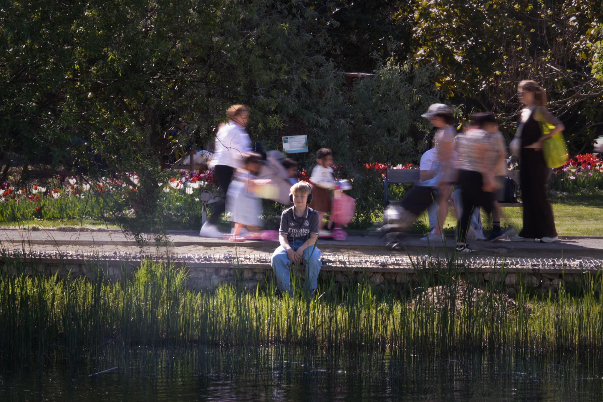 Eine Person sitzt allein an einem Teich und blickt in die Kamera, während eine Gruppe von Menschen, die im Hintergrund vorbeigeht, verschwommen erscheint, wodurch in der Parkkulisse ein Kontrast zwischen Stille und Bewegung entsteht.