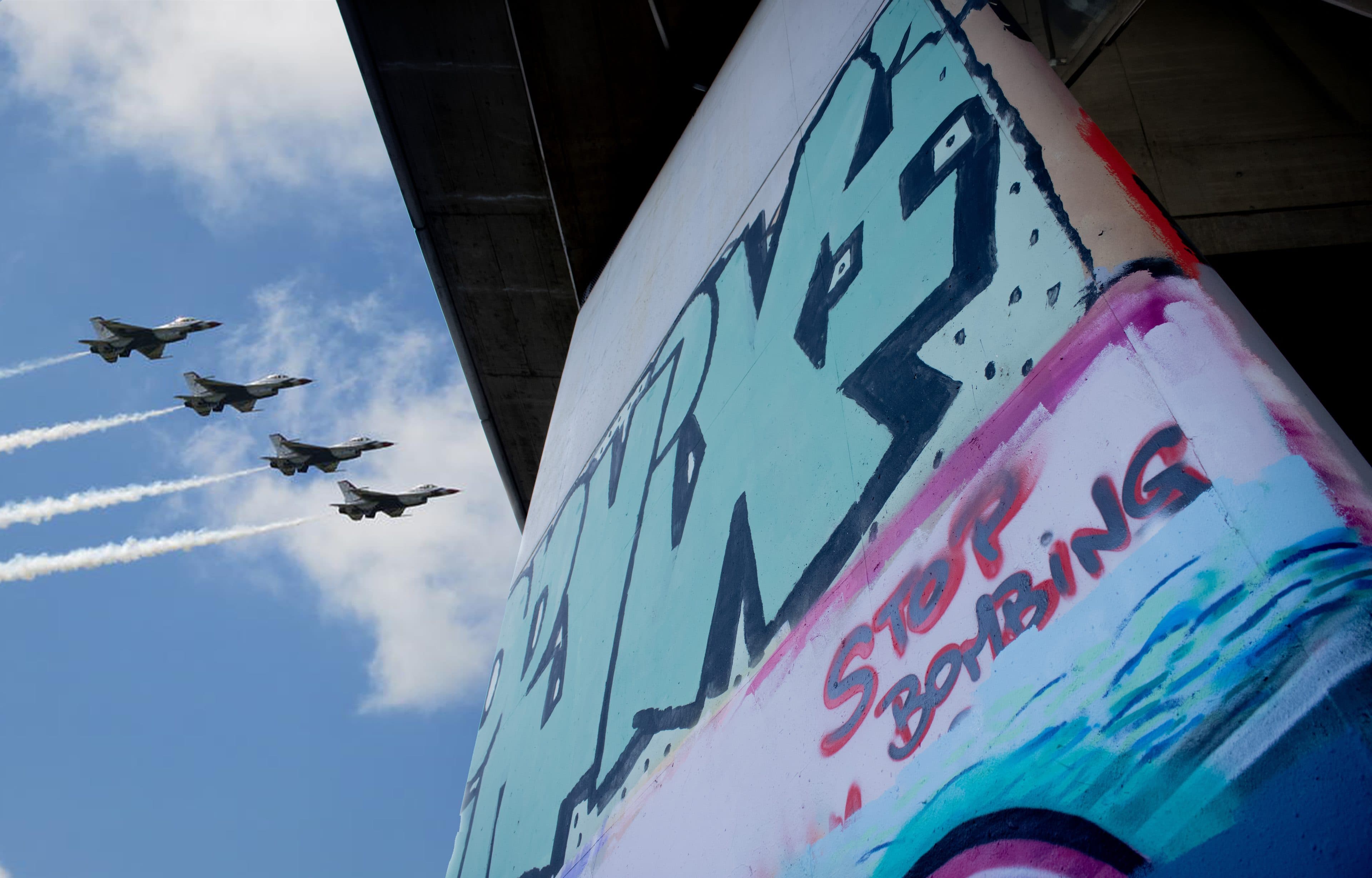Vier Kampfjets fliegen in Formation und ziehen weiße Kondensstreifen am blauen Himmel hinter sich her. Im Vordergrund ist auf einer Gebäudesäule ein buntes Graffiti zu sehen, auf dem in roter Farbe die Worte „STOP BOMBING“ gesprüht sind.