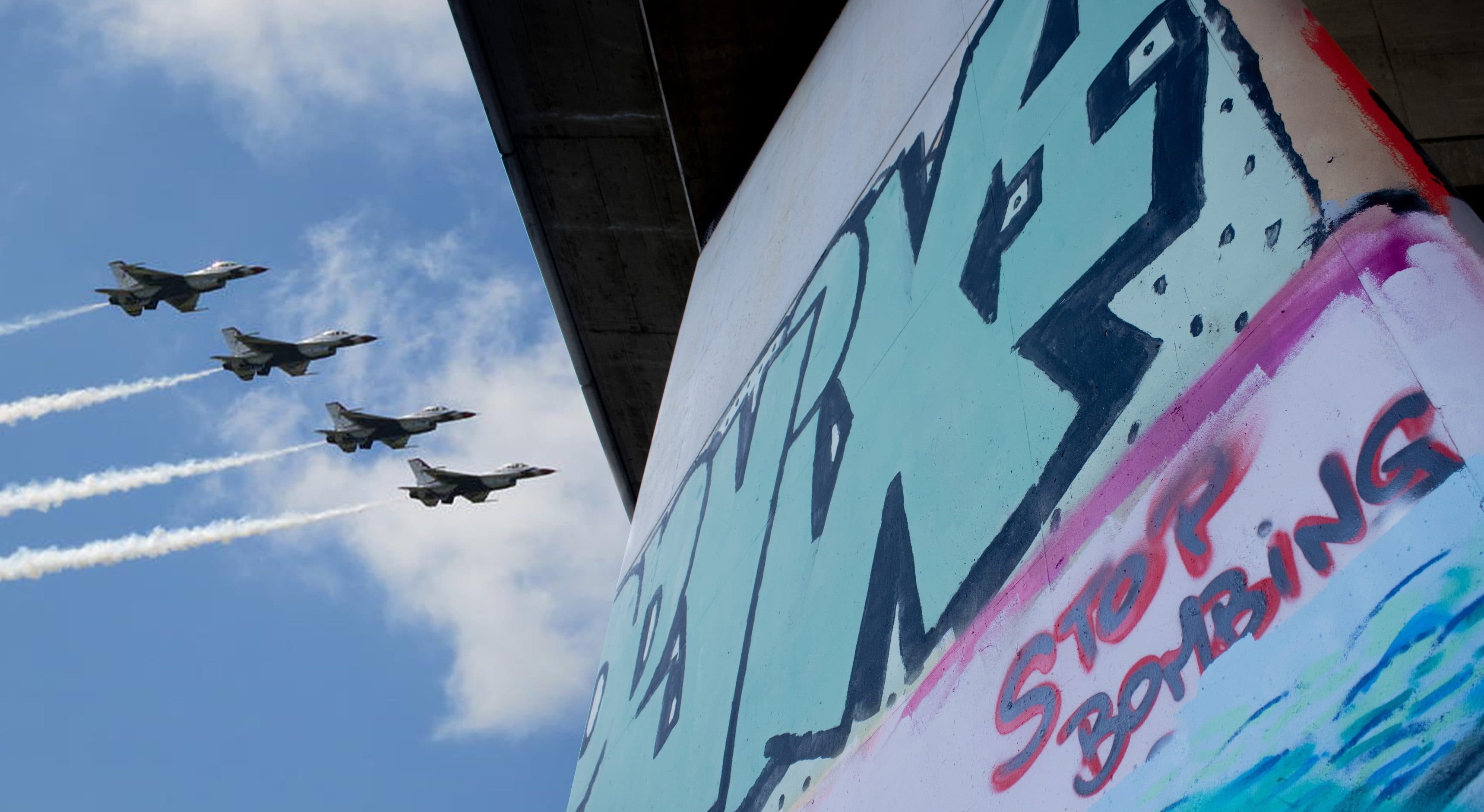 Vier Kampfflugzeuge fliegen in Formation und ziehen weiße Kondensstreifen am Himmel hinter sich her, in der Nähe eines Brückenpfeilers, der mit bunten Graffiti-Schriftzügen bedeckt ist, darunter die Worte „STOP BOMBING“. Die Szene vereint Elemente aus Militär und Straßenkunst.