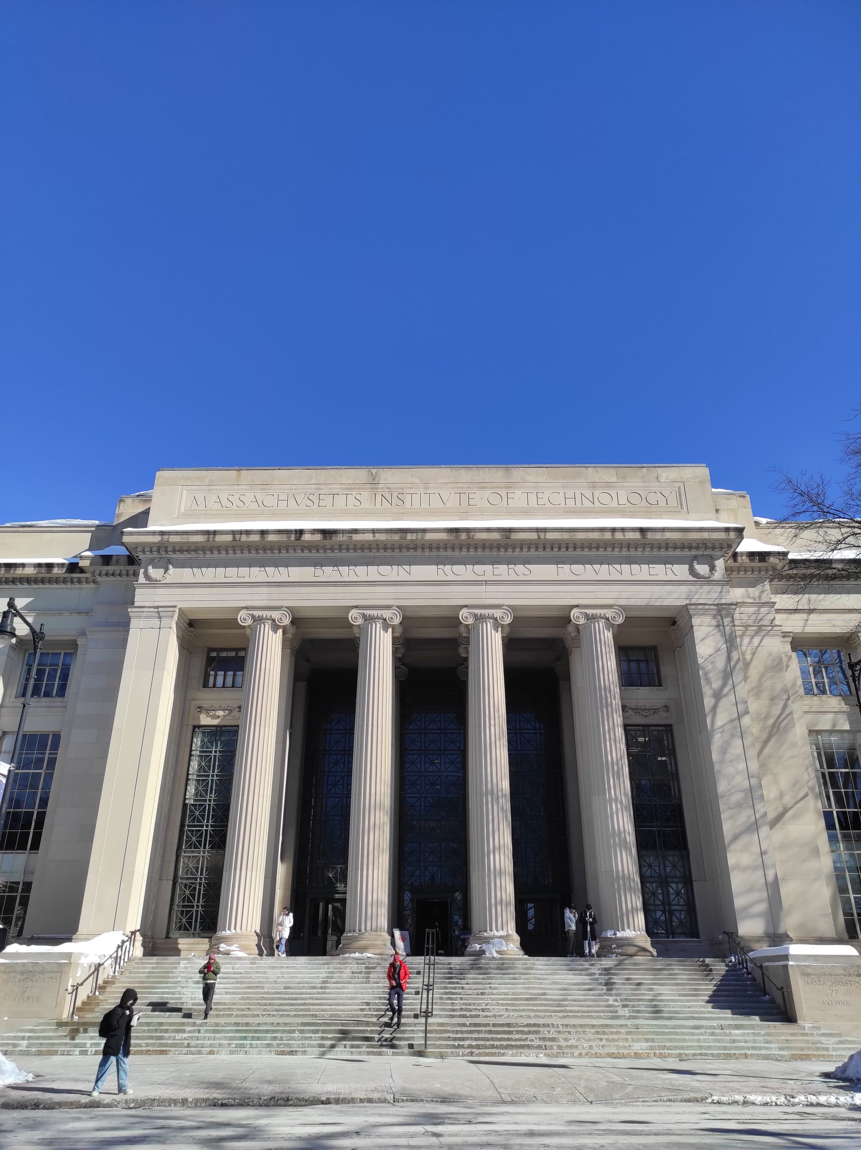 Die Fassade des Massachusetts Institute of Technology (MIT) mit hohen Säulen, großen Glastüren und Menschen, die unter strahlend blauem Himmel die Stufen hinaufgehen.
